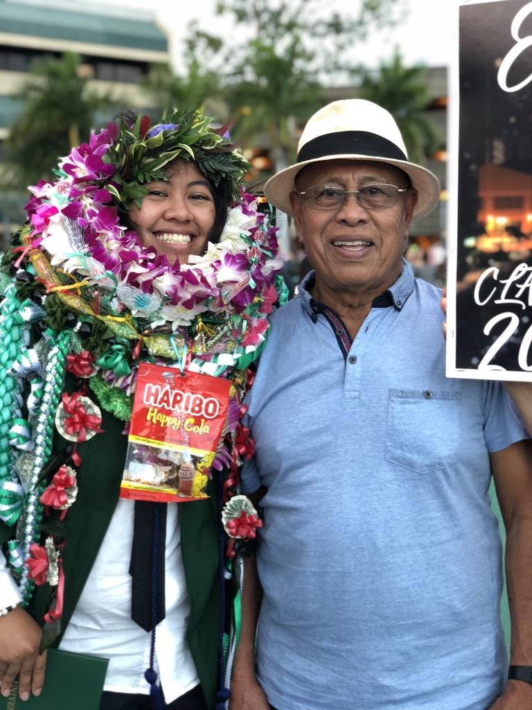 A happy graduate stands with a proud elder, both smiling widely amidst festive decorations.
