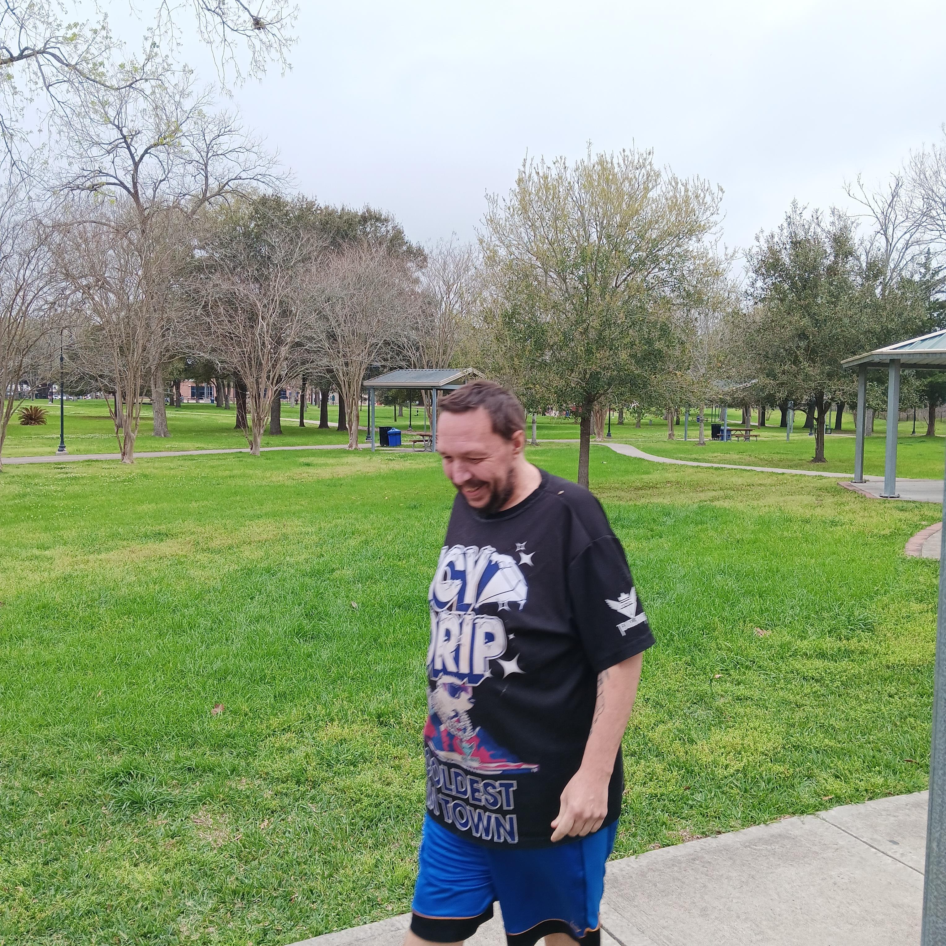 A man in casual wear strolls through a vibrant park with trees and picnic spots on a cloudy day.