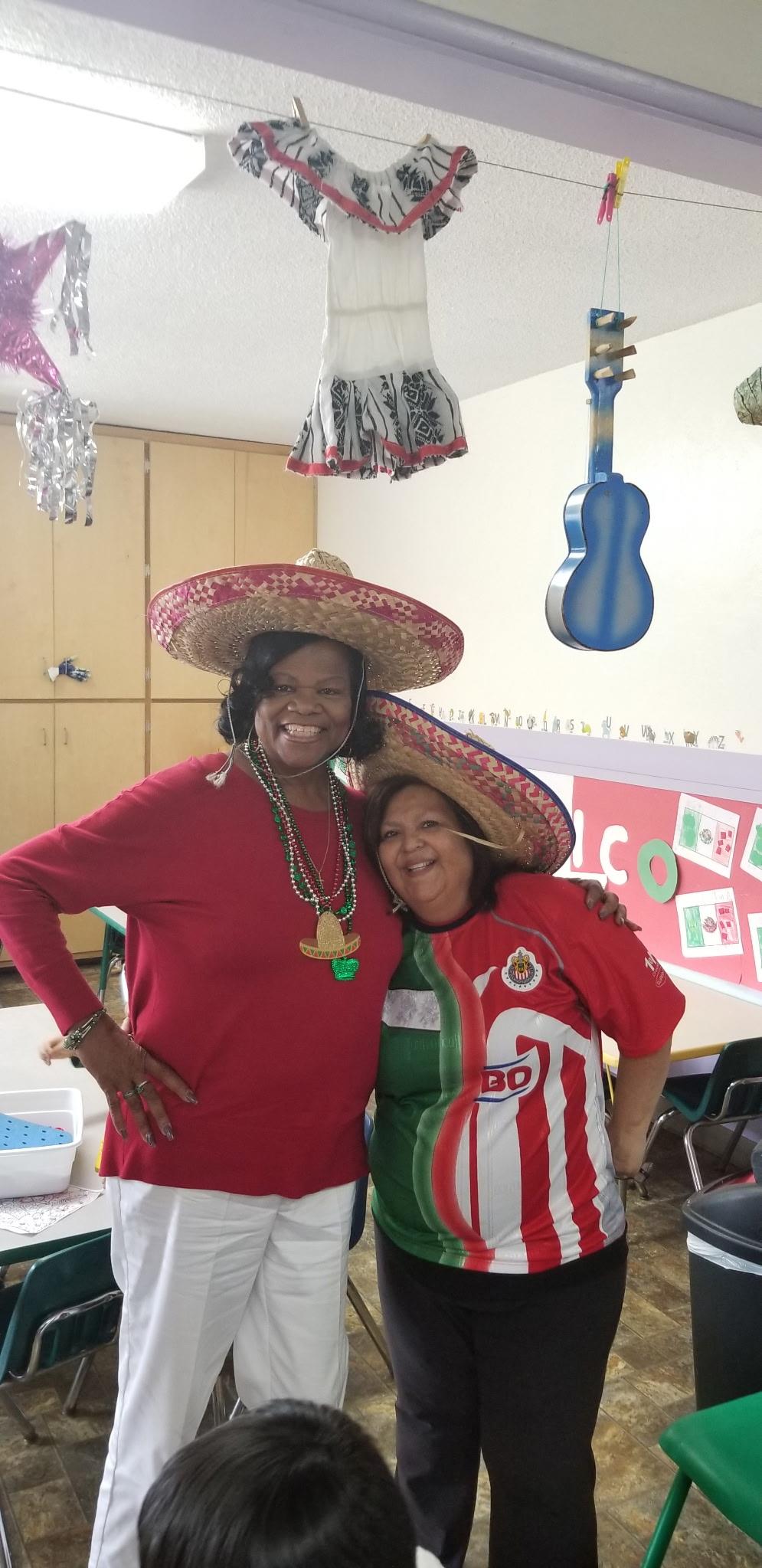 Two women wear traditional hats and smile while celebrating in a colorful classroom setting.