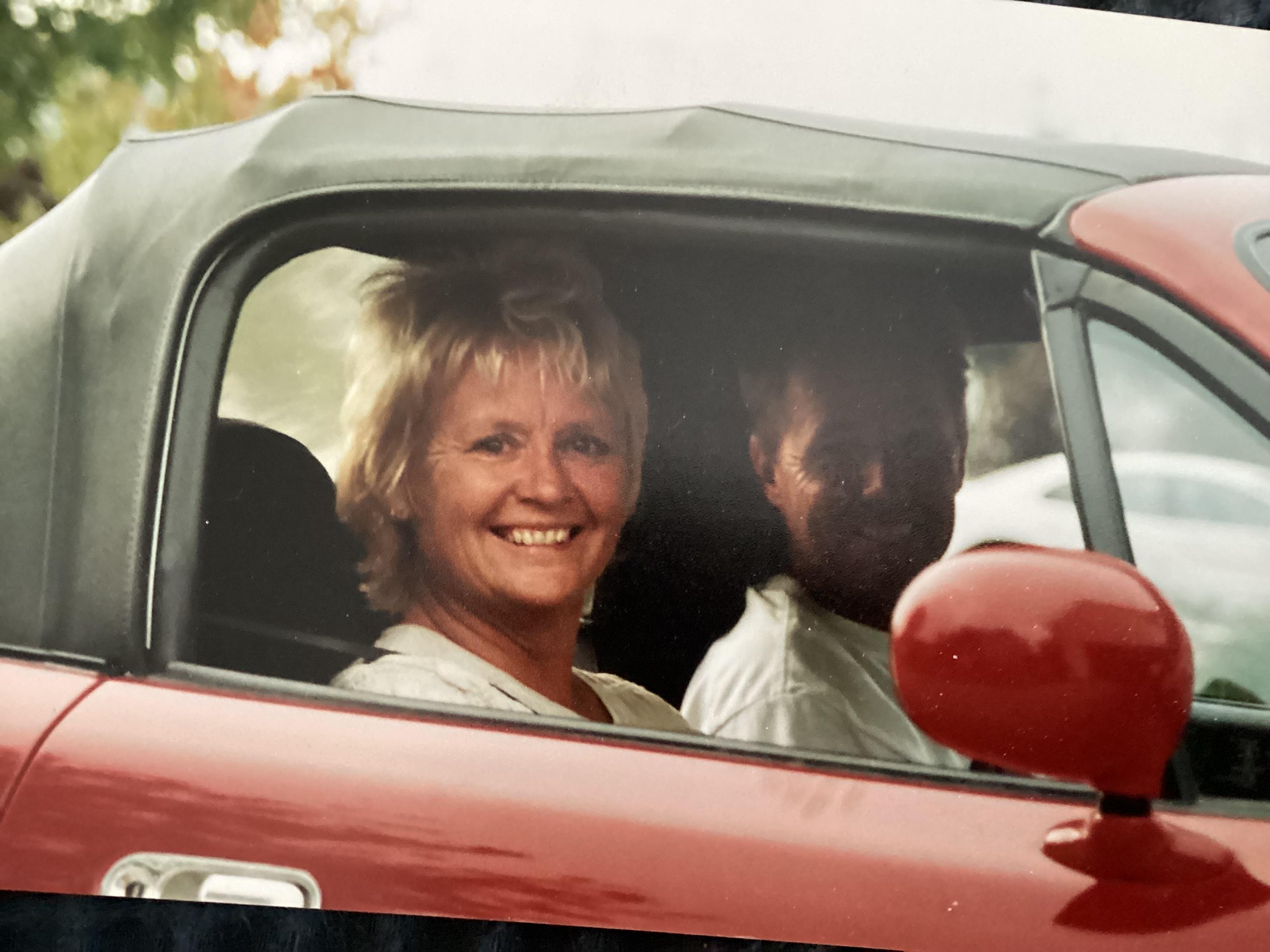 Two friends smile happily while sitting in a red convertible under a clear sky.