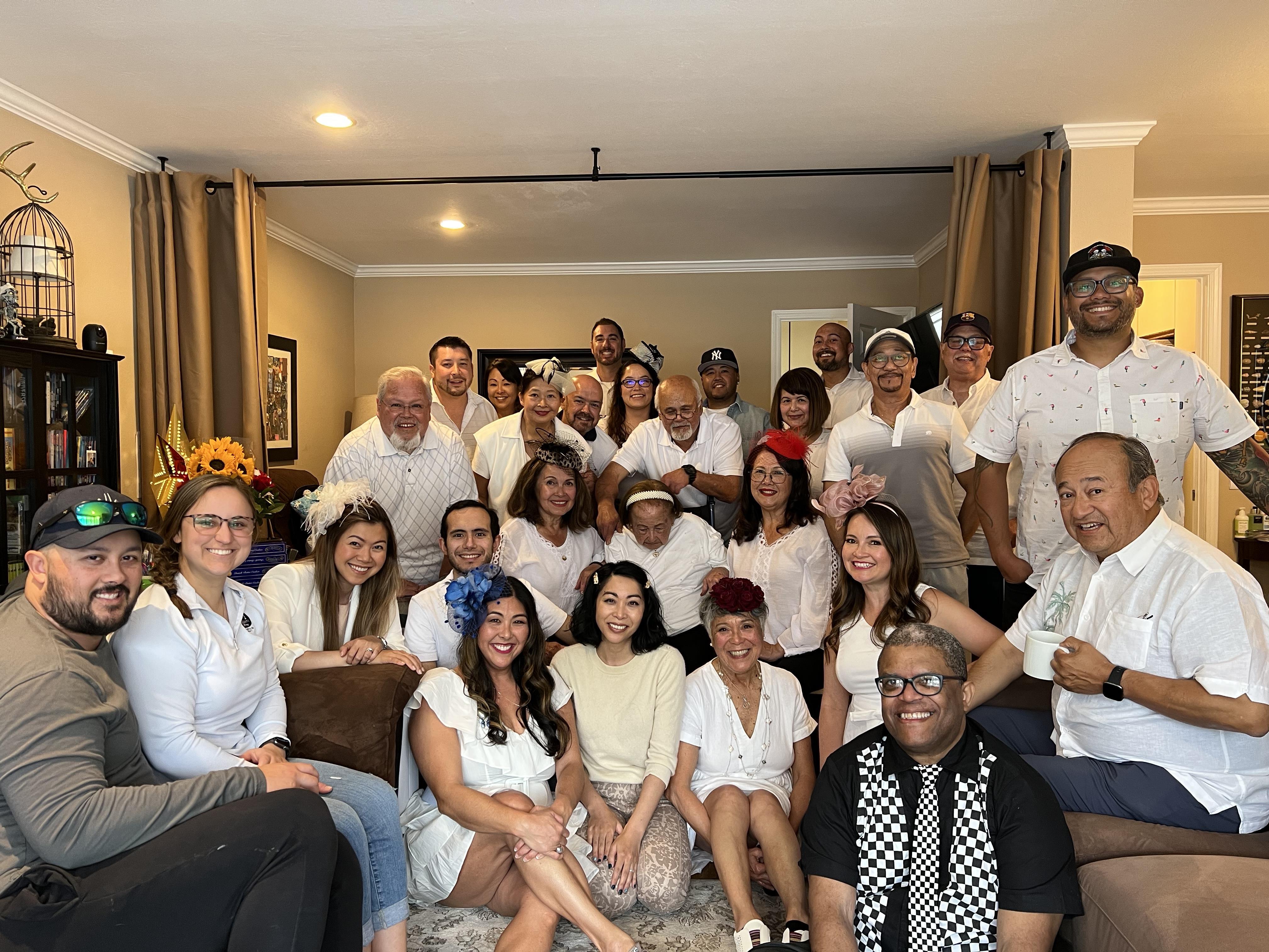 A large group of friends and family members dressed in white celebrate together inside a warm home.