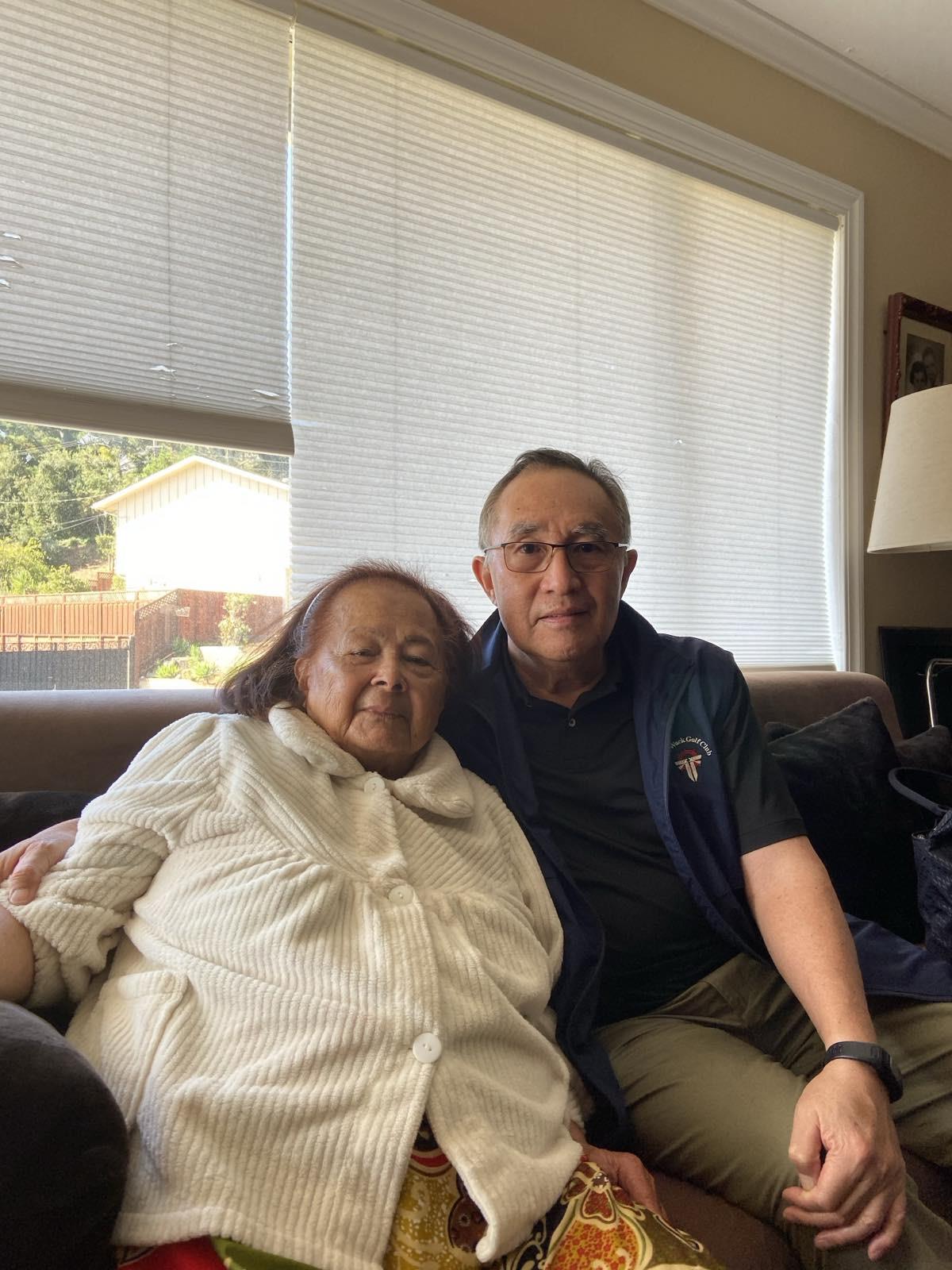 A younger man sits next to an elderly woman, both smiling, in a cozy living room.