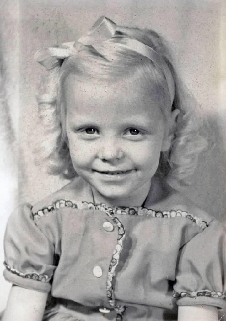 A cheerful young girl with blond curls and a bow sits for a portrait, wearing a vintage dress.