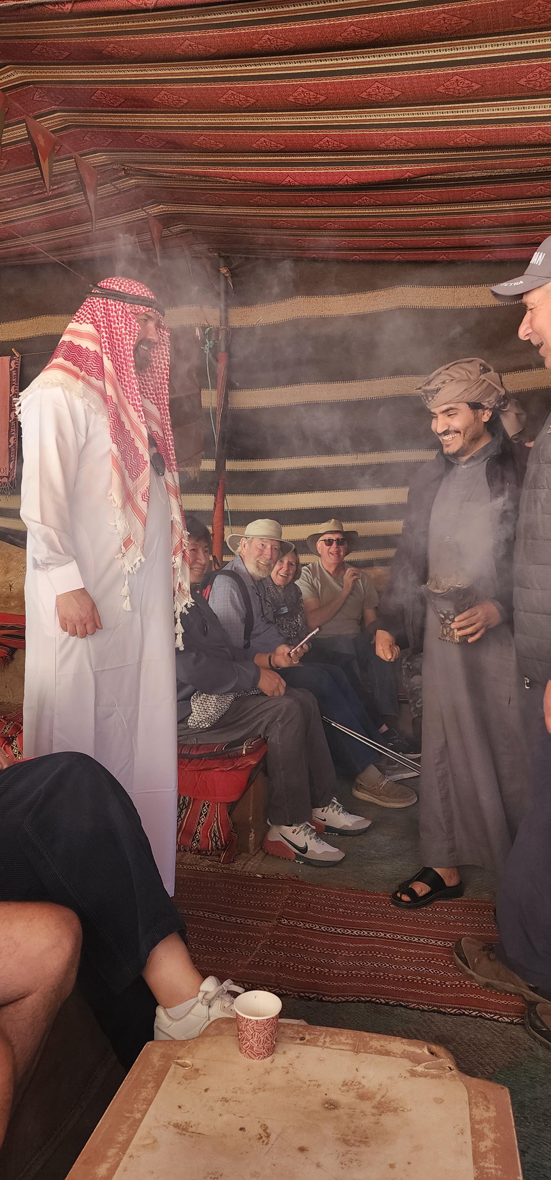 Men engage in conversation and share drinks inside a cozy desert tent filled with smoke.