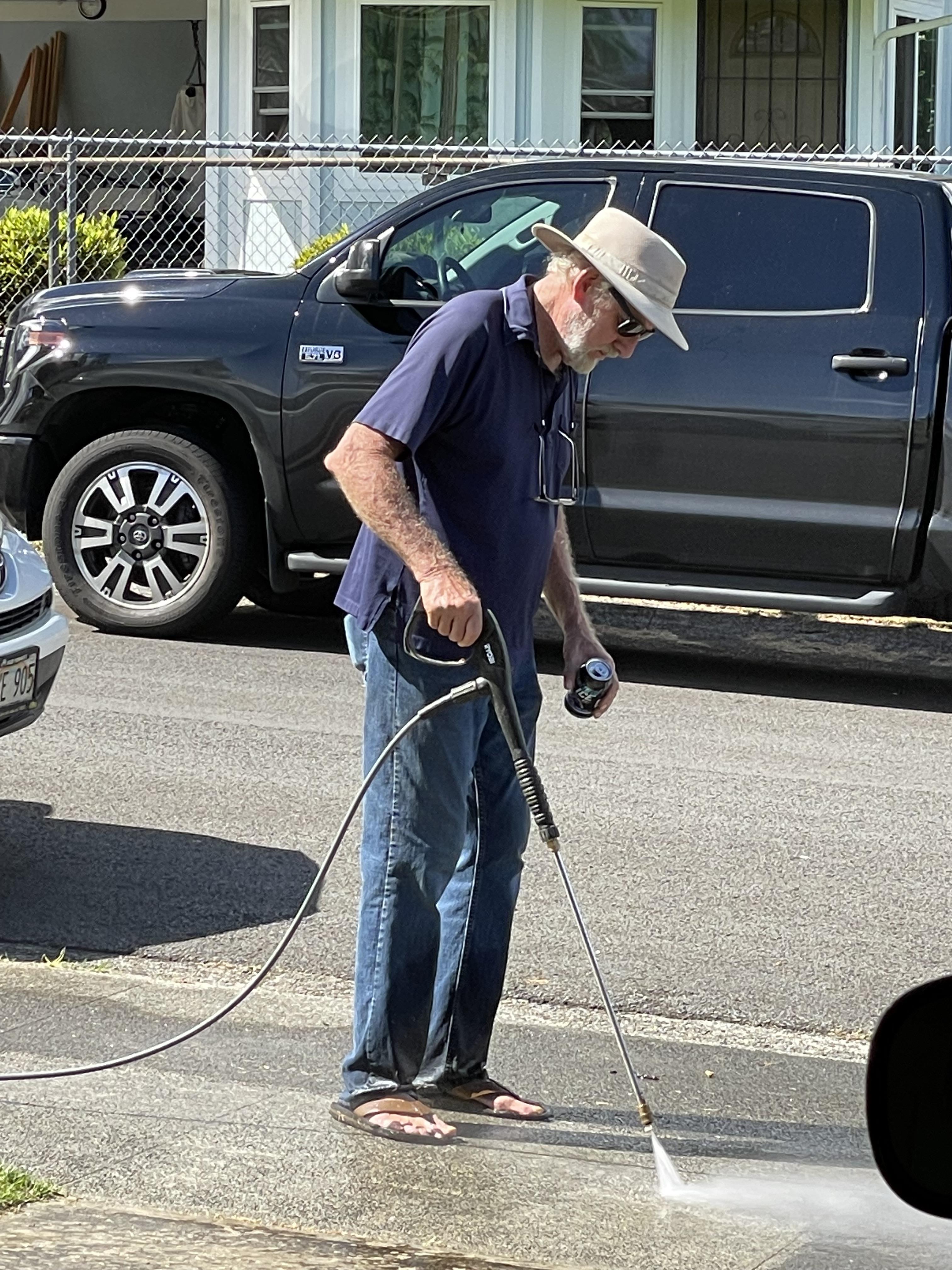 Senior man cleans his driveway using a pressure washer while wearing casual clothes and sunglasses.