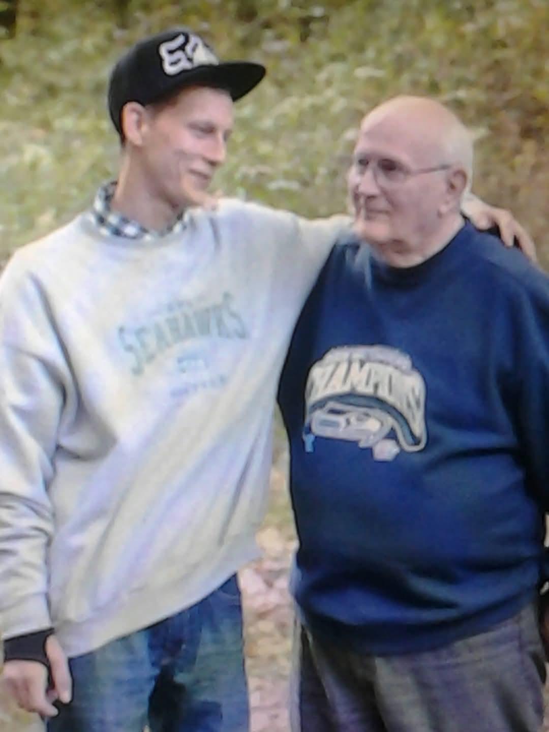 Two men stand close, smiling and enjoying a warm conversation in nature.
