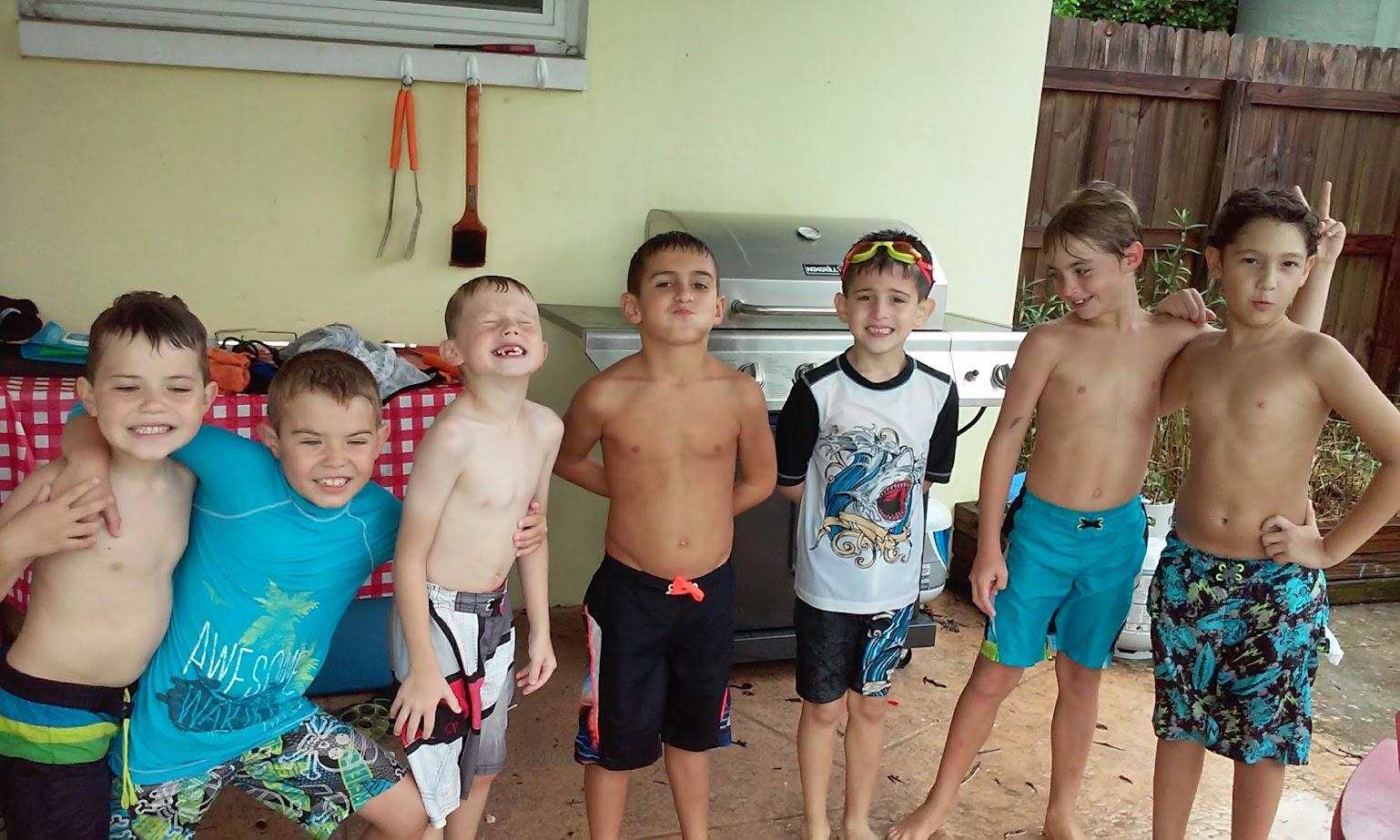 Seven boys gather smiling near a grill, wearing swimwear and enjoying a fun summer day outdoors.