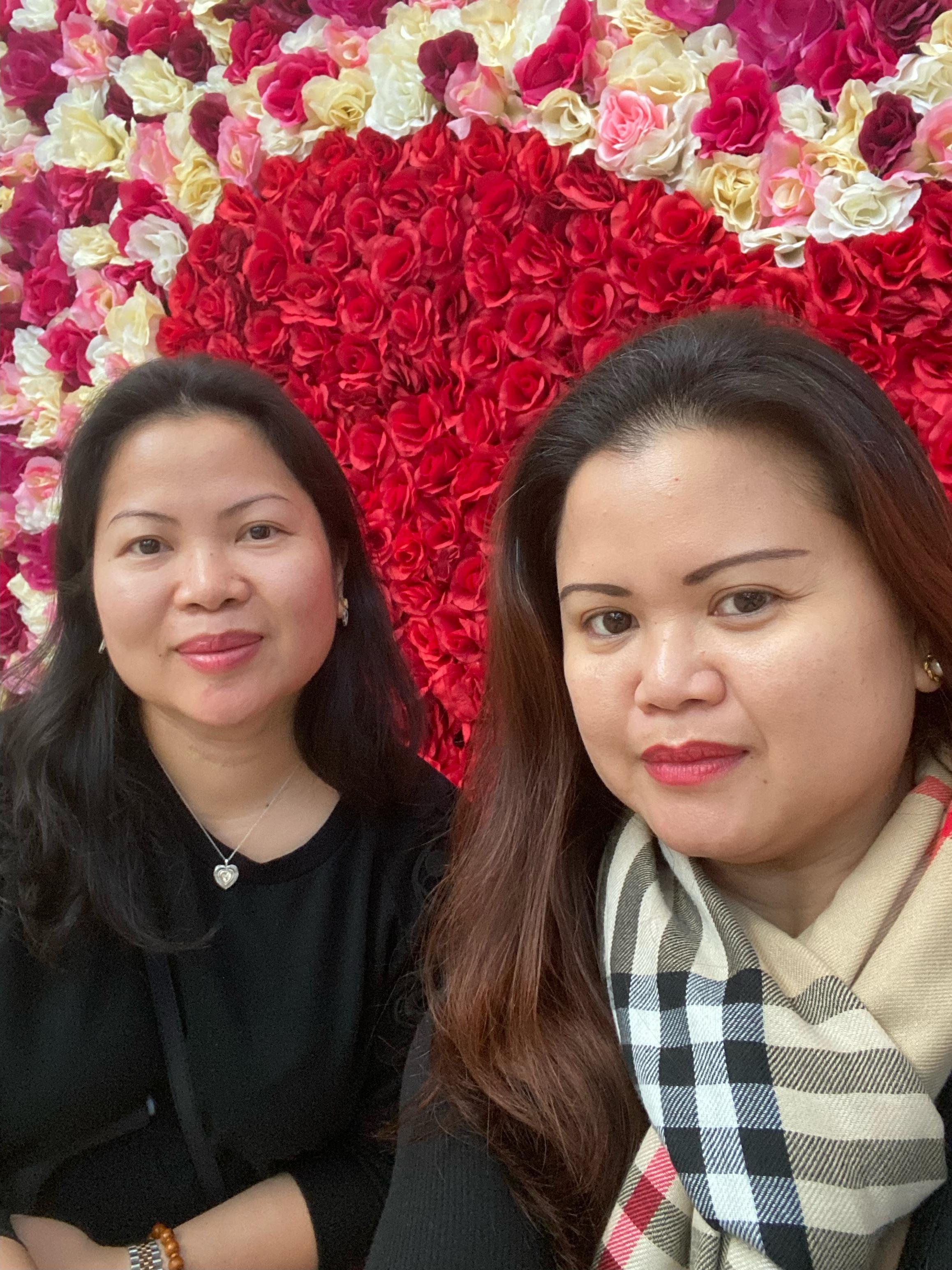 Two women smile for a selfie against a backdrop of red and pink flowers at an event.