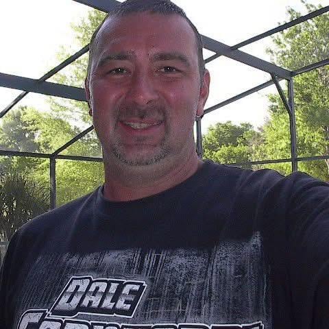 A man stands in a sunny screened porch smiling with trees visible outside in the background.
