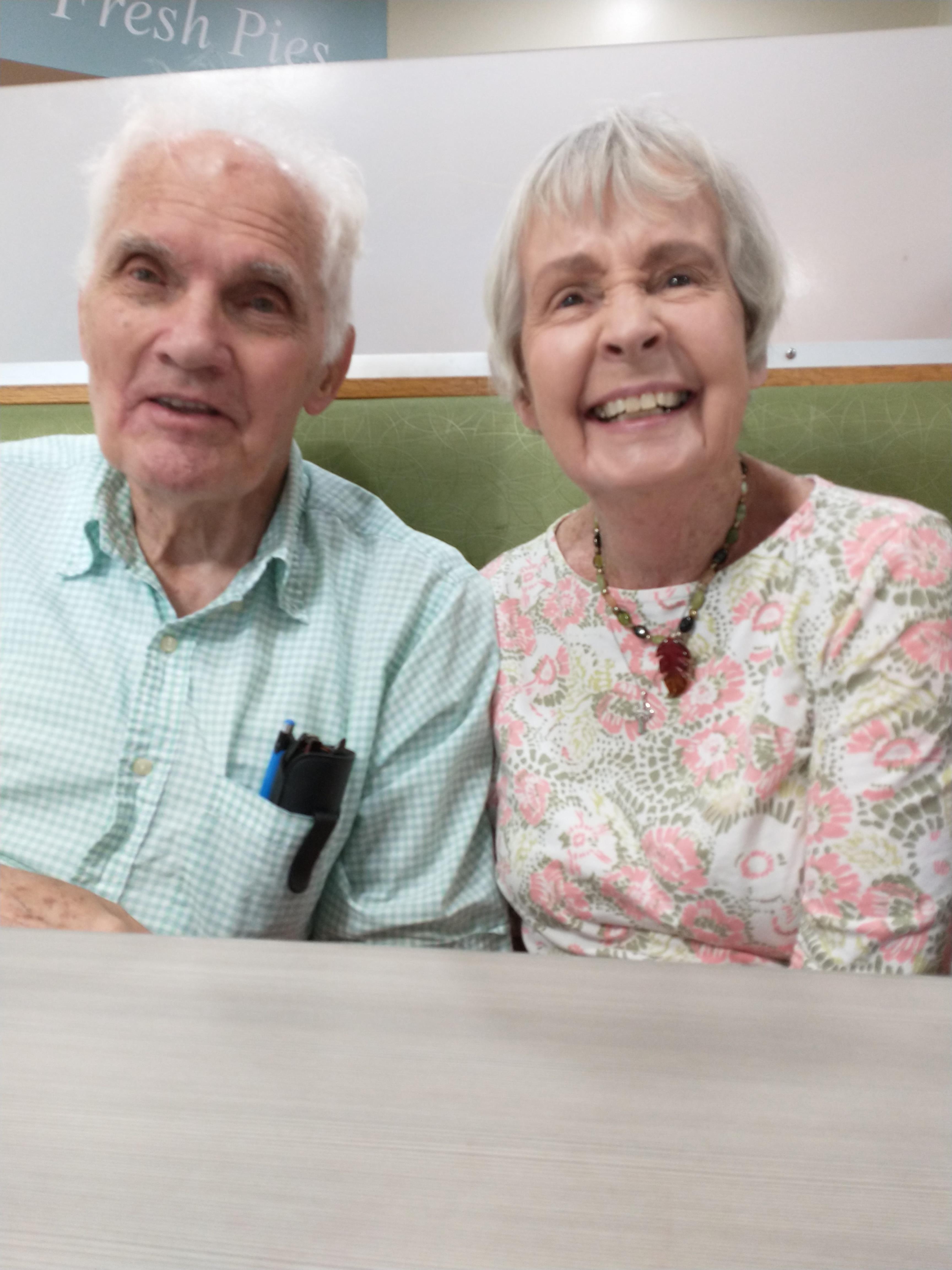 Two smiling seniors sit closely together at a table, sharing happiness in a bright environment.