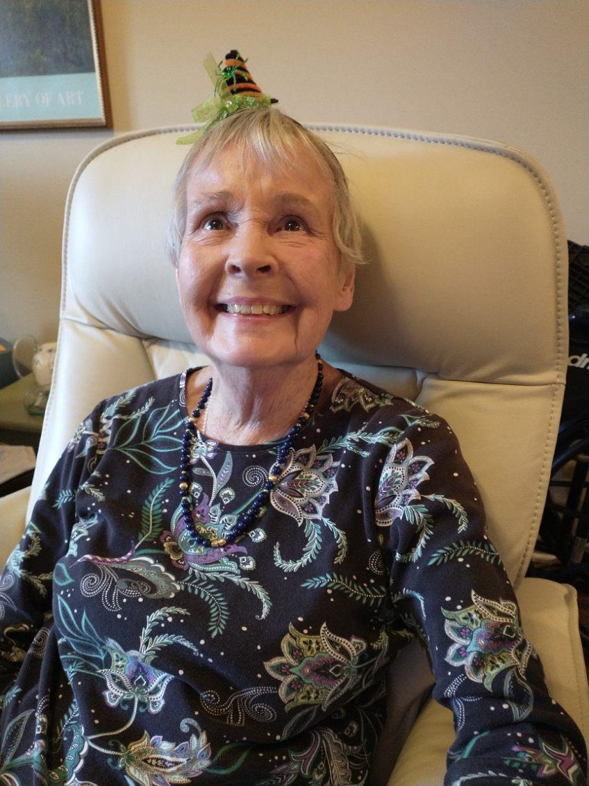 A smiling elderly woman with a colorful hair accessory sits in a cozy chair.