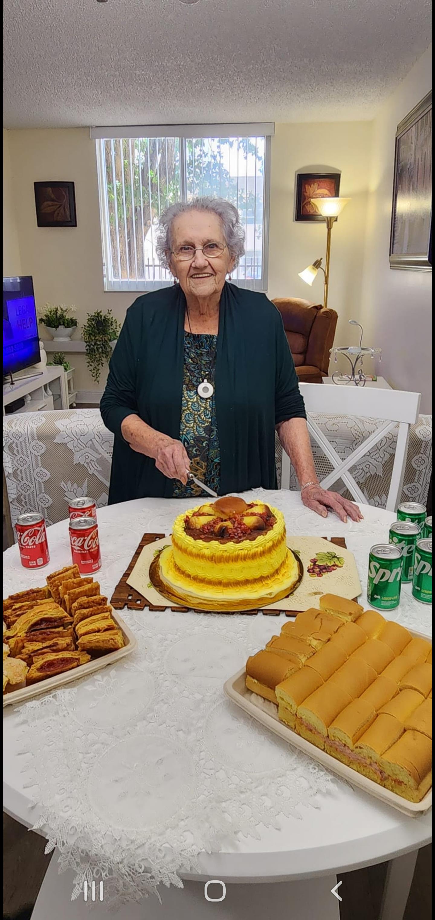 Elderly woman smiles while cutting a yellow cake surrounded by various snacks and beverages.