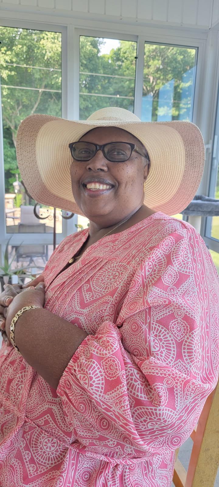 A cheerful woman smiles while wearing a stylish sun hat and sitting comfortably inside a sunroom.