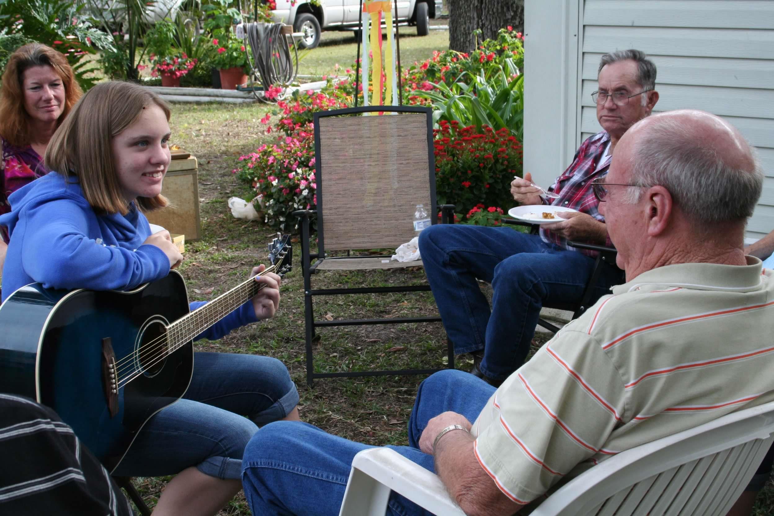 A girl strums her guitar while relatives enjoy food and engage in conversation in the yard.