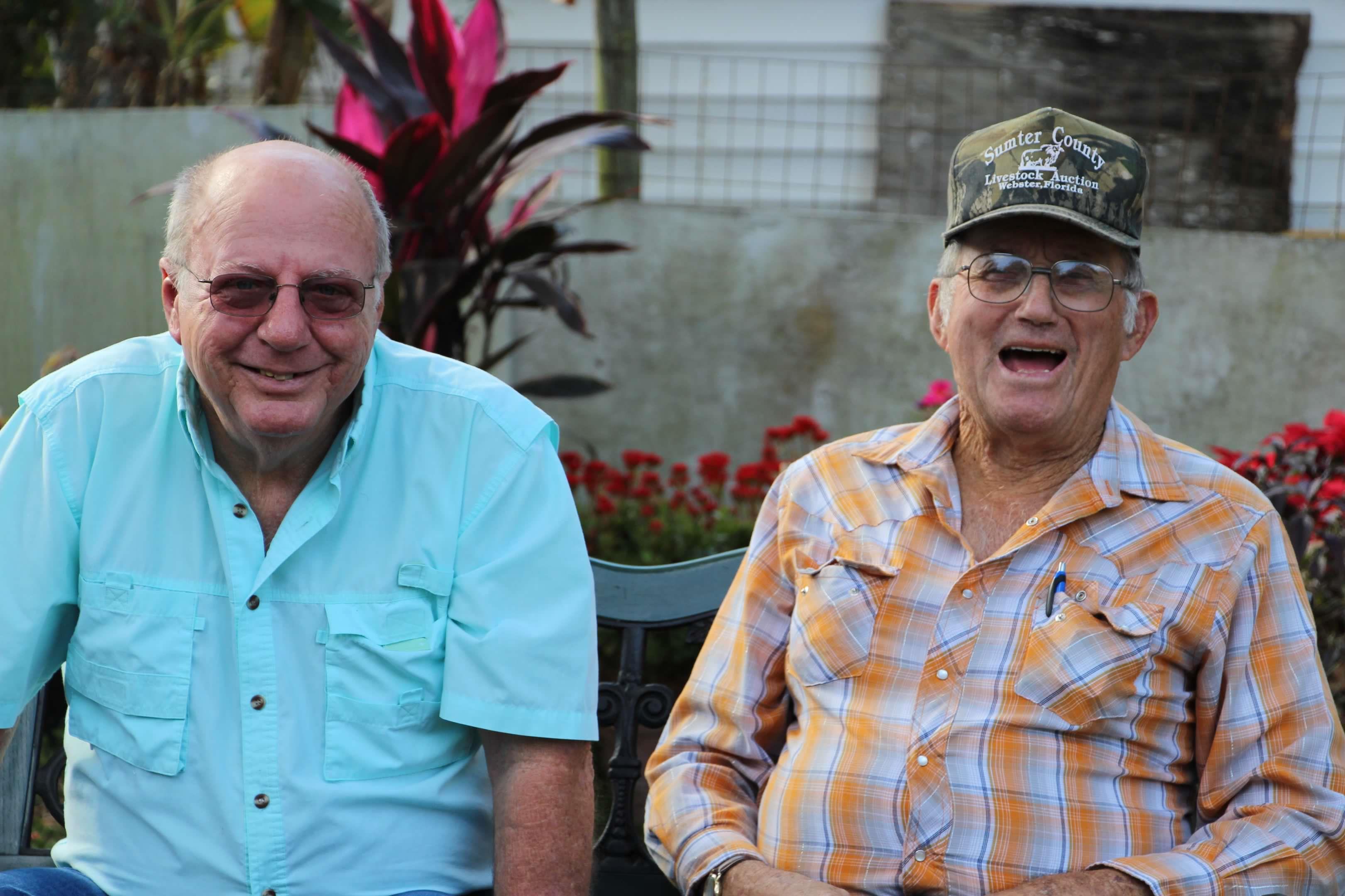 Two friends share laughter while sitting in a colorful garden filled with flowers and greenery.