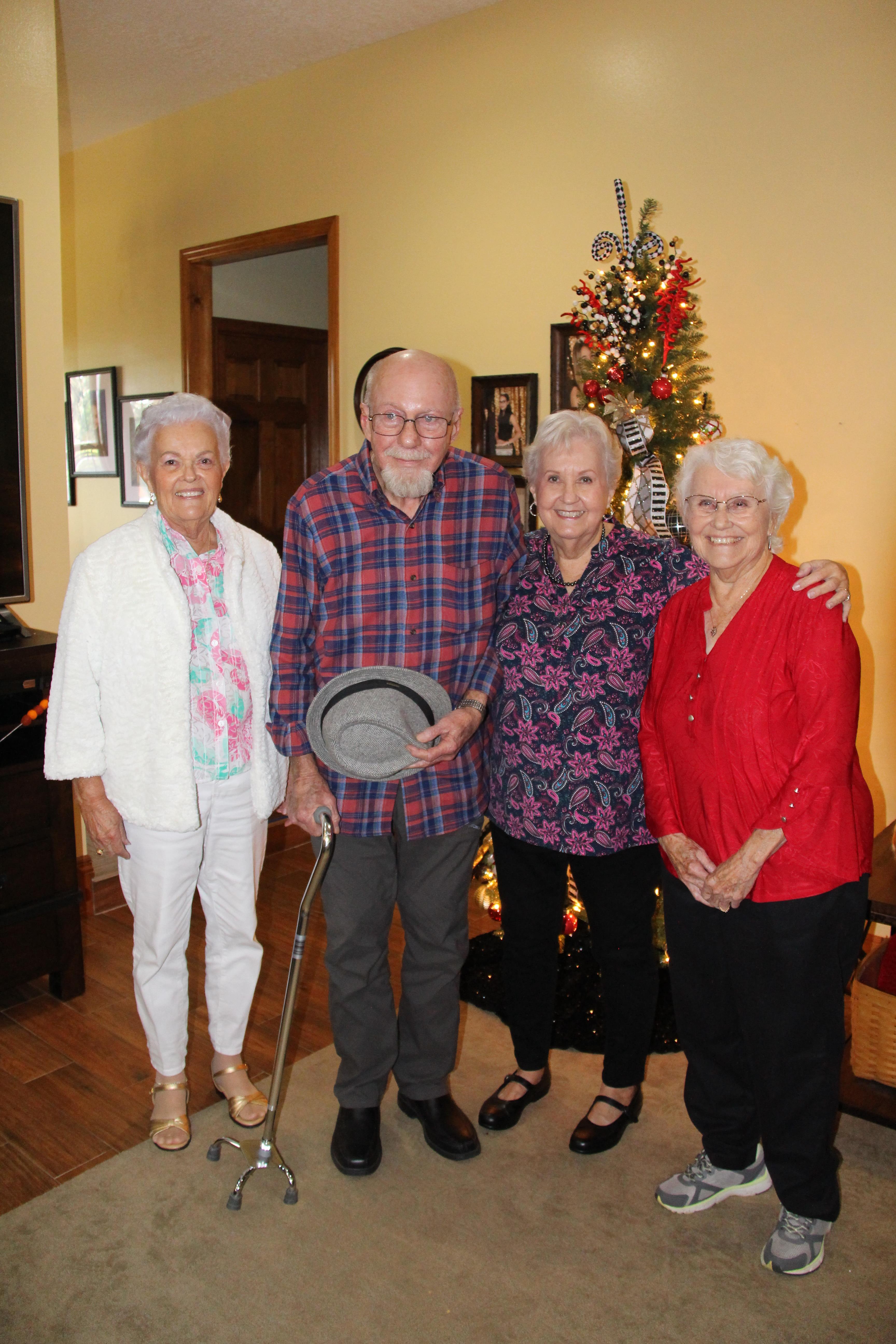 Four friends gather and smile joyfully in front of a decorated Christmas tree at home.