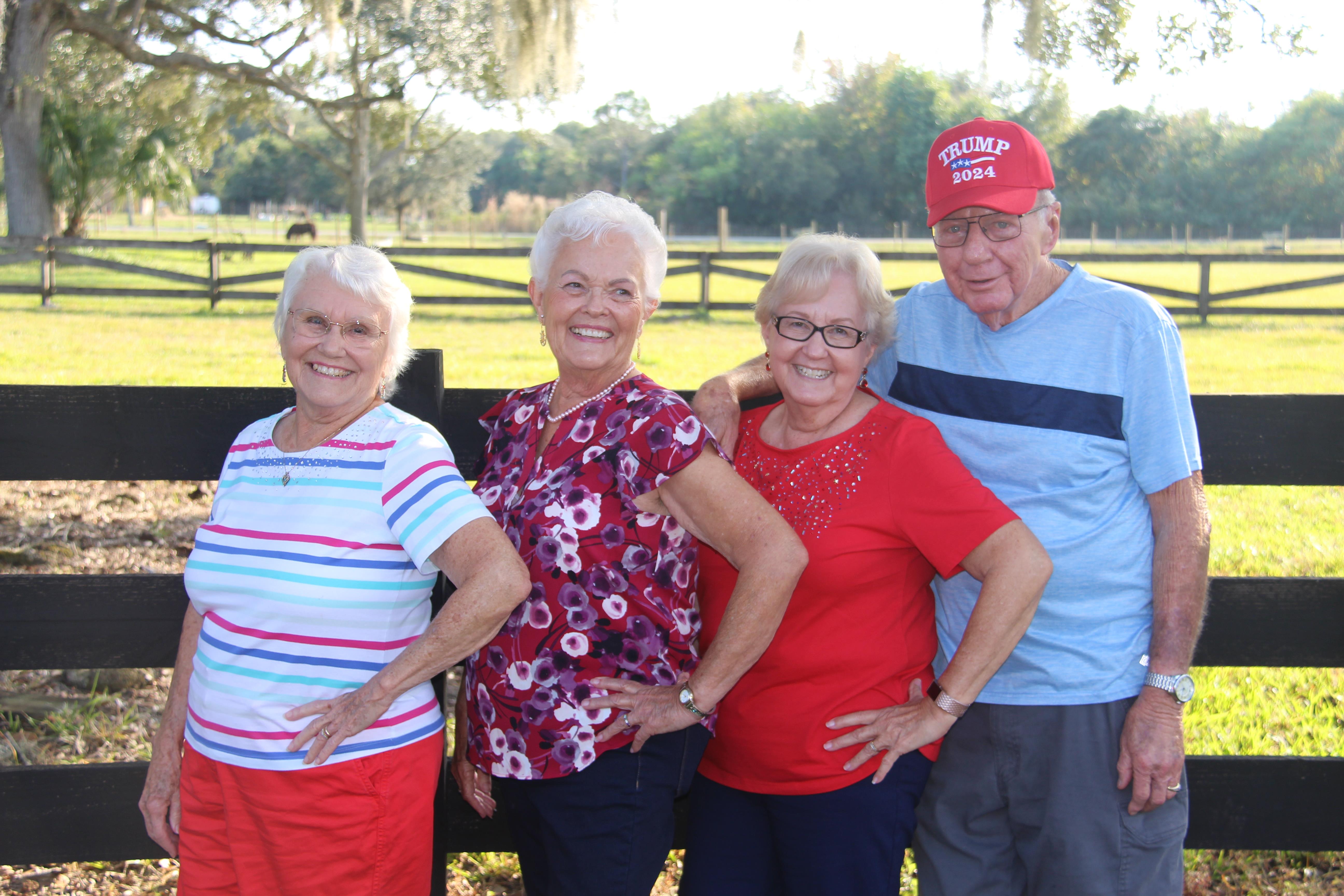 Four friends enjoy time together outdoors, posing happily against a wooden fence in bright sunlight.