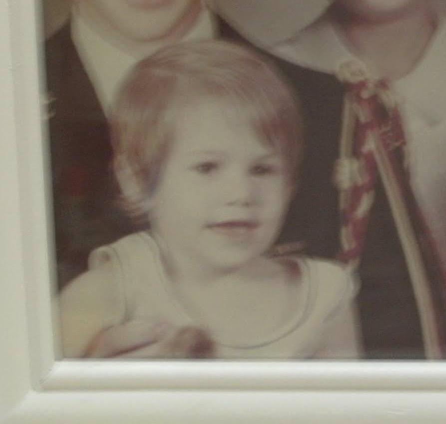 A young child smiles at the camera, standing with adults in a cozy setting.
