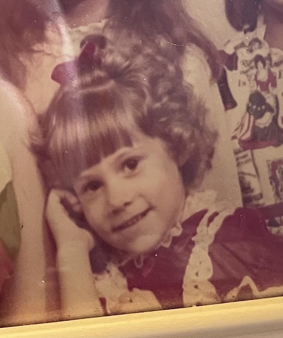 A young girl in a festive dress smiles happily, surrounded by friends in a warm home setting.