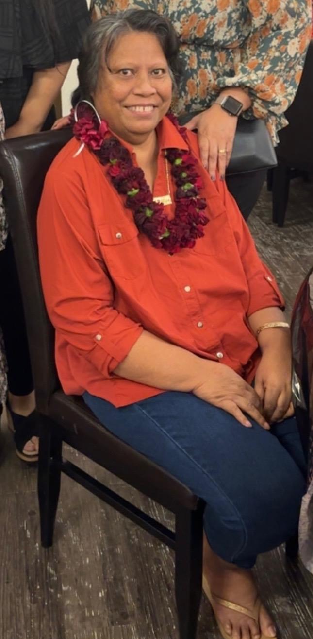 Woman in a red shirt and flower lei sits smiling in a relaxed indoor setting with friends.