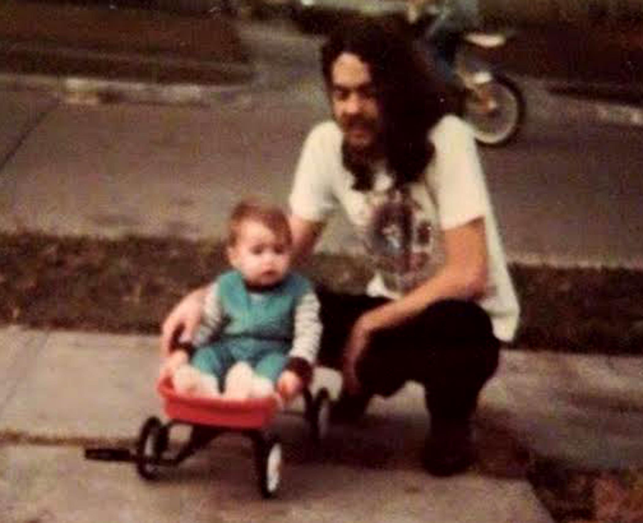 A man kneels next to a child in a red wagon, surrounded by a sunny suburban setting.