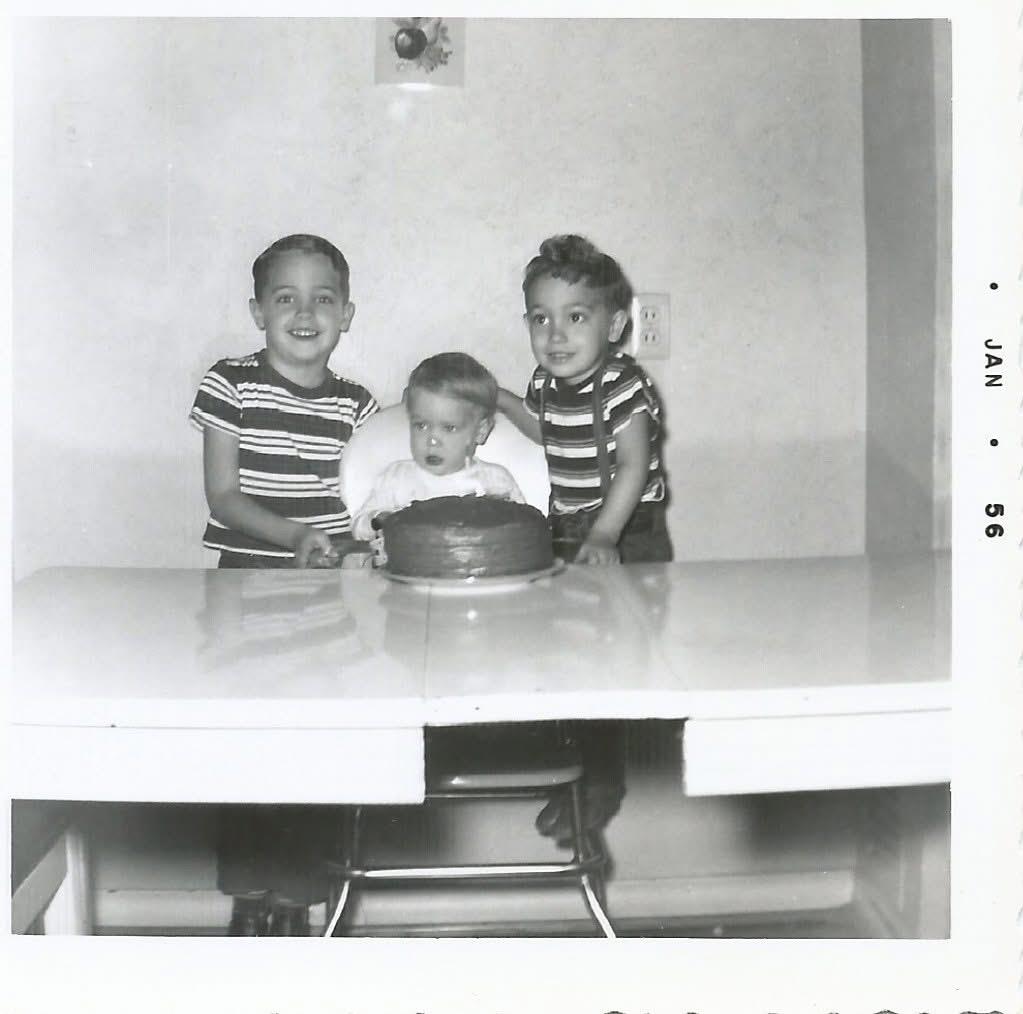 Three young boys gather around a cake, smiling joyfully in a bright indoor space.