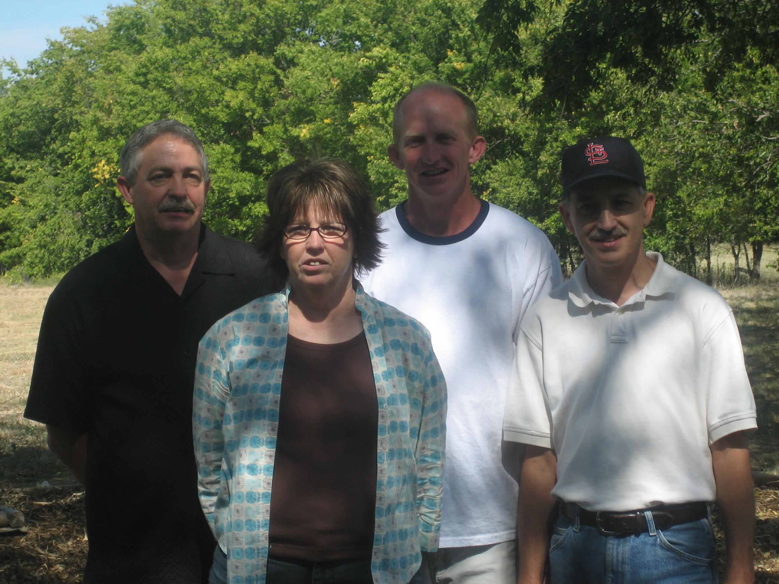 Four individuals stand close together in a sunny park, enjoying a warm afternoon outdoors.