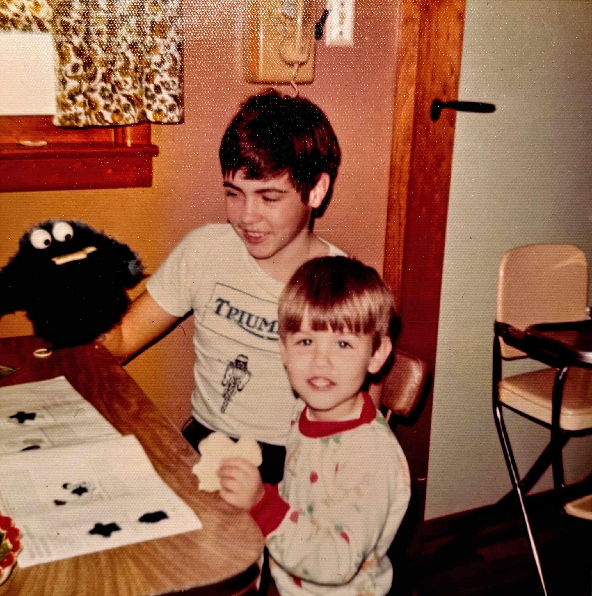 Two boys engage in a creative activity at a table, one holding a puppet while snacking.