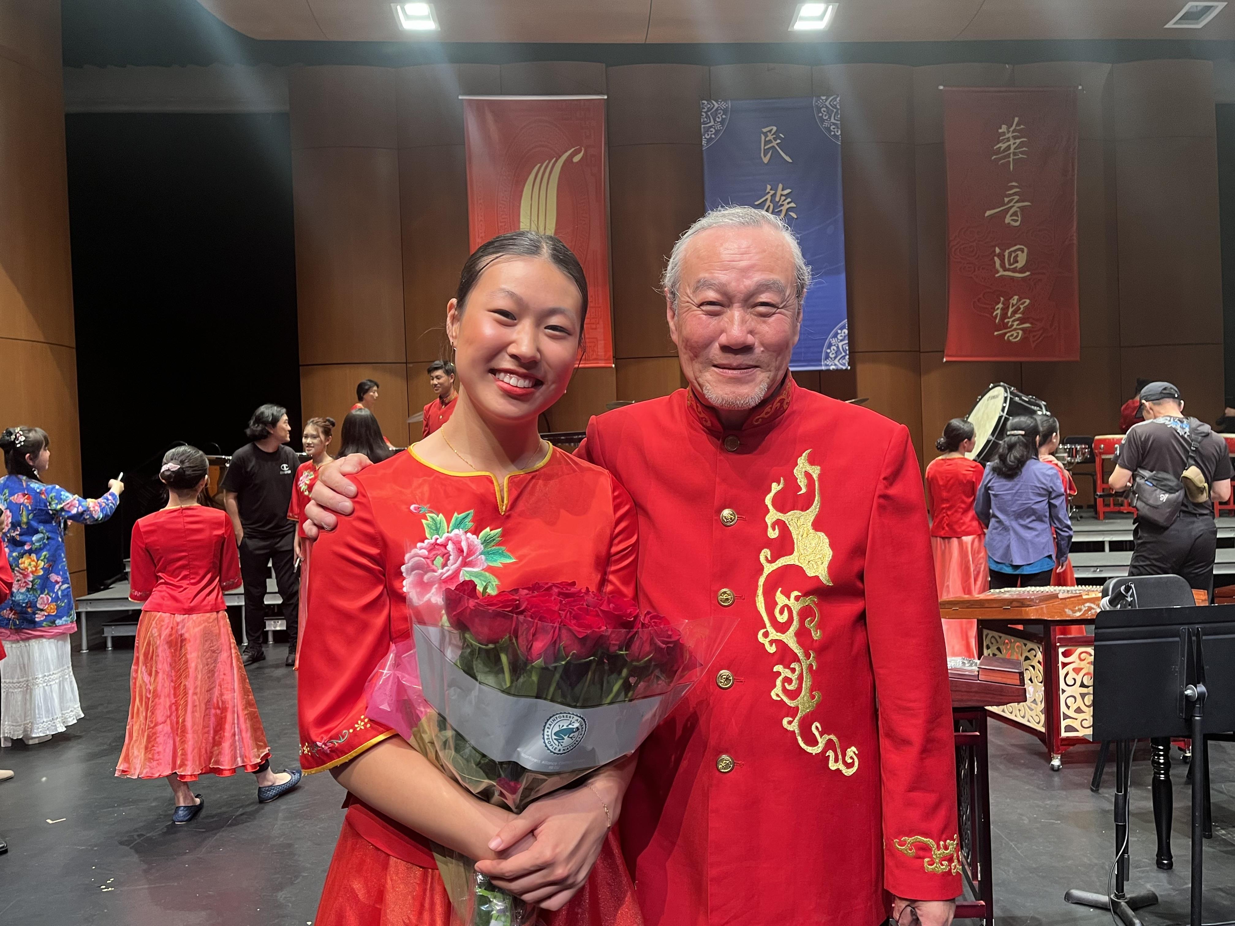 A performer dressed in red holds flowers while posing with an elder during a cultural event.