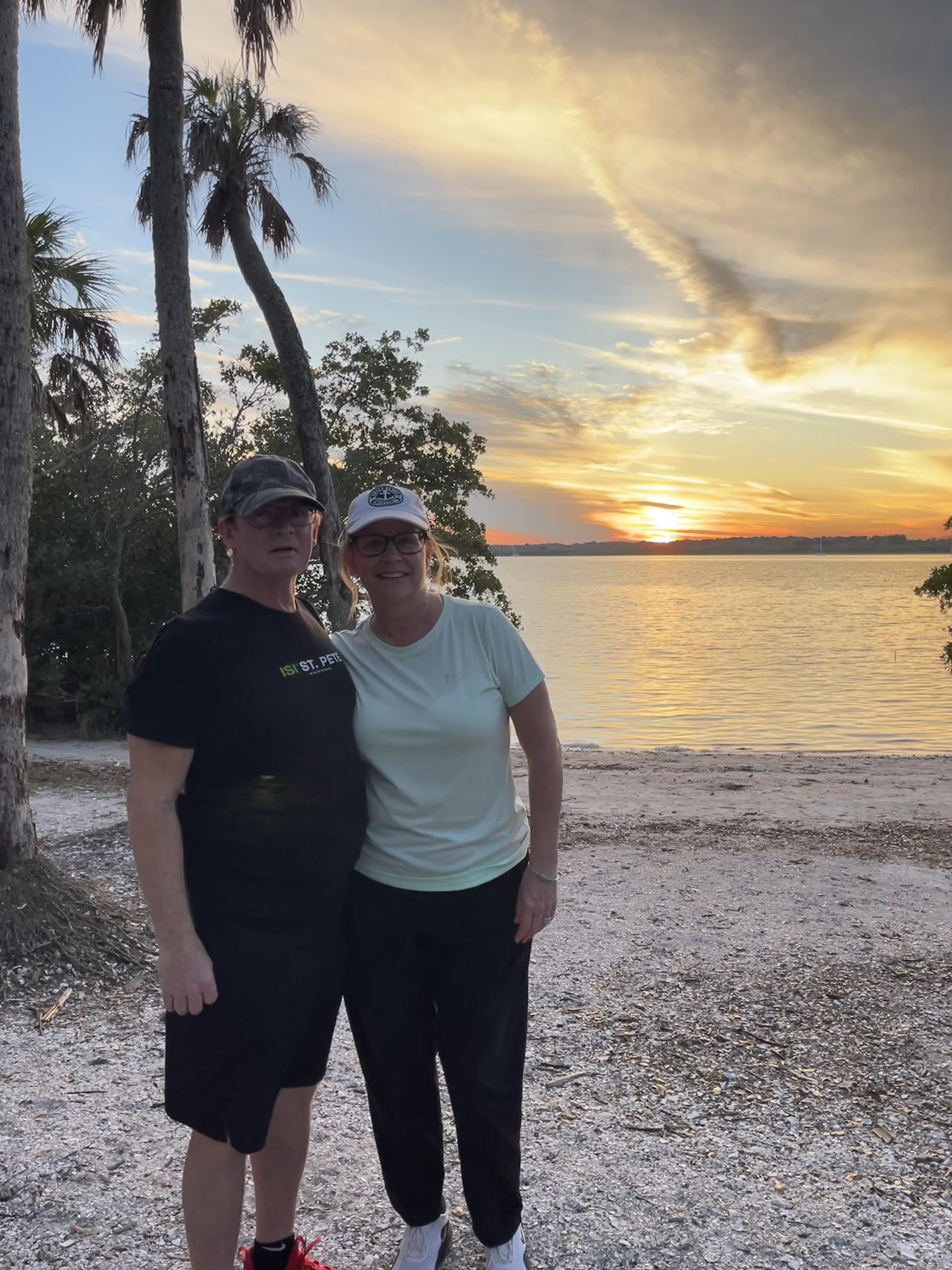 Two friends pose together at a beach during sunset, enjoying the beautiful scenery and company.