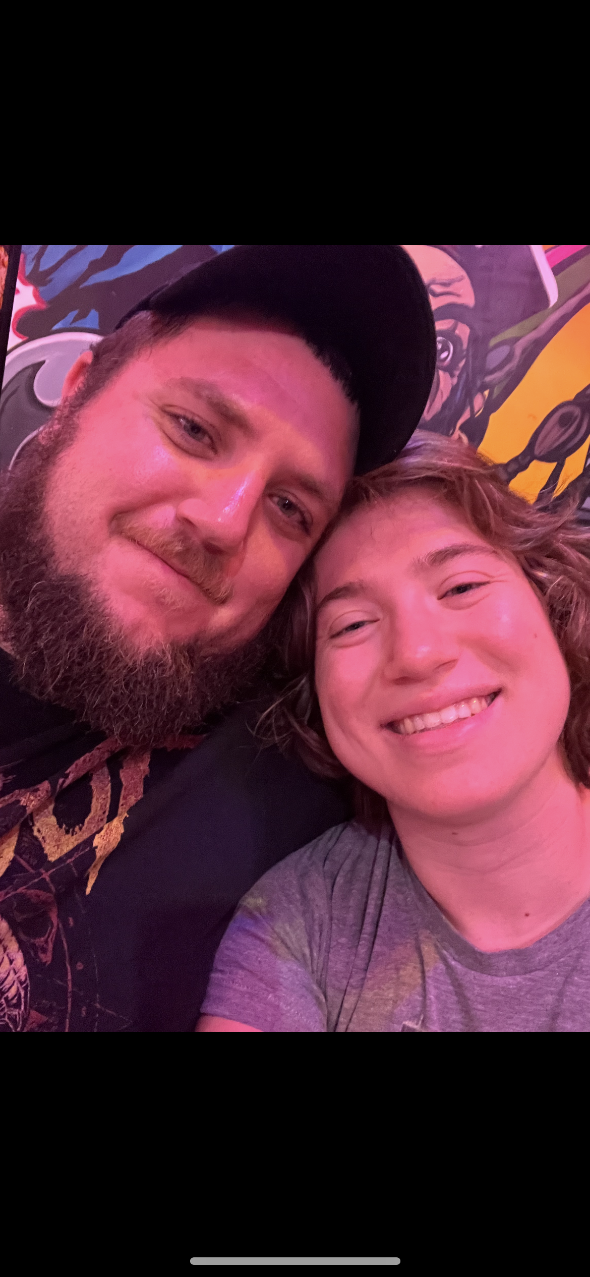Two friends smile widely while sitting close together in front of vibrant street art.