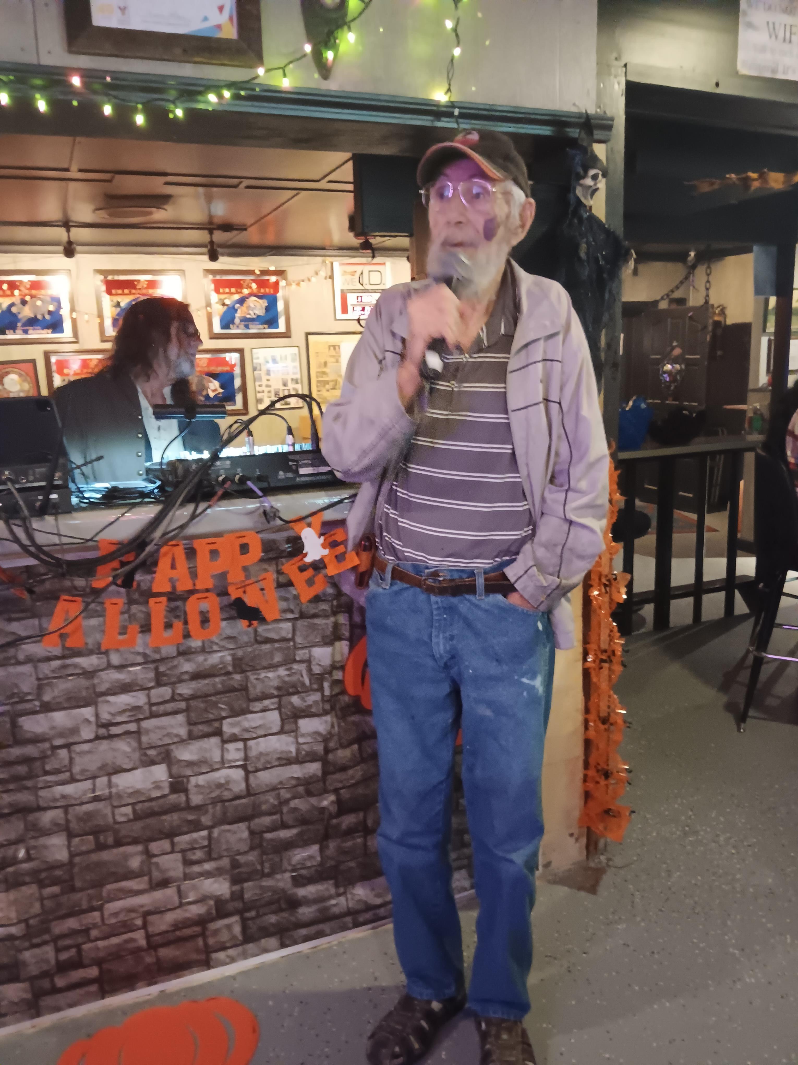 An older man sings karaoke in a festive bar while a band plays in the background.