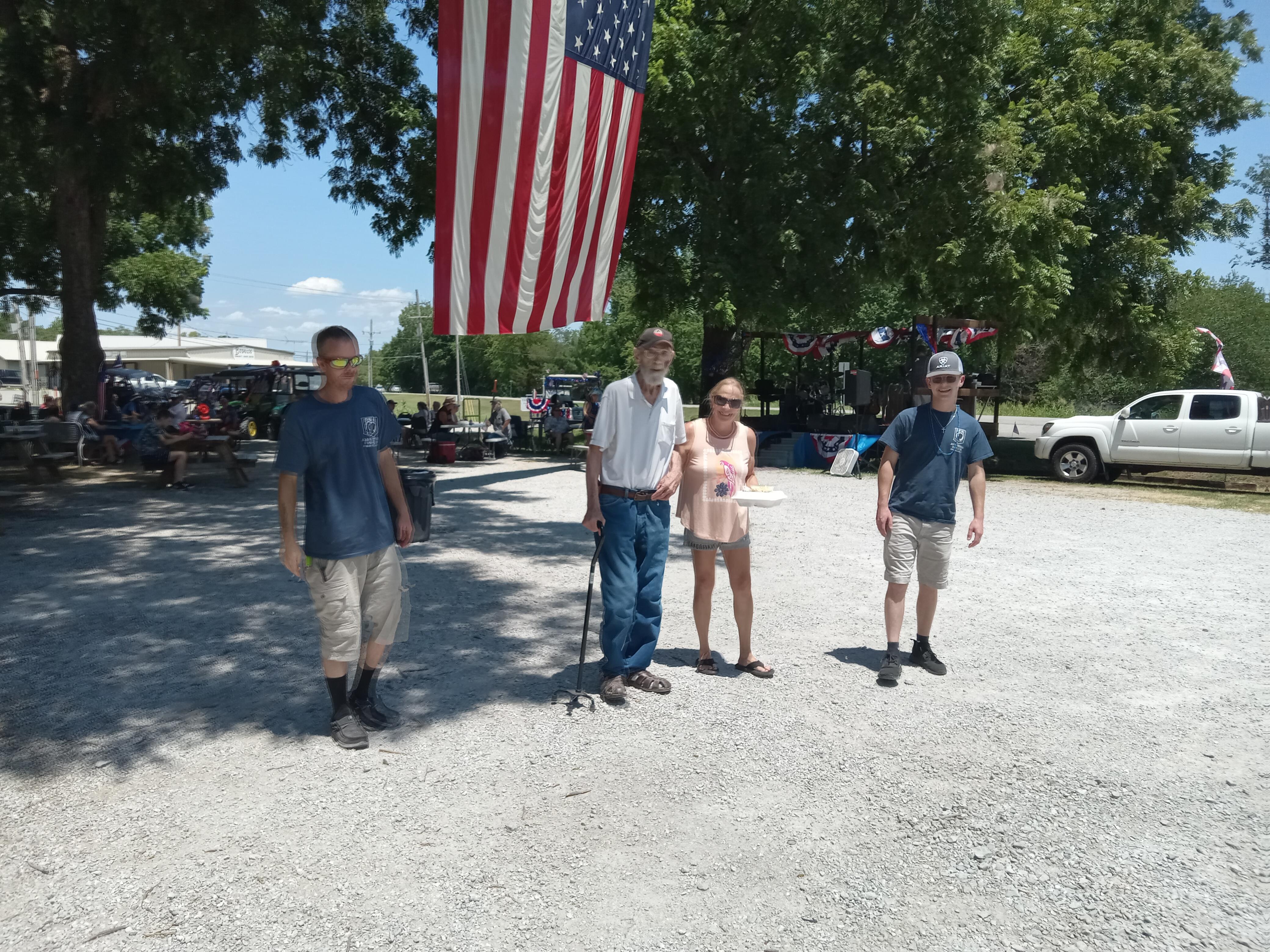 Group of four individuals stands near a giant American flag in a sunny outdoor area.