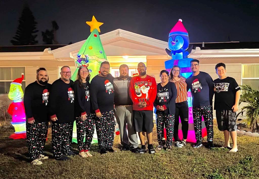 Group of family members poses happily in festive attire by their brightly lit holiday decorations.