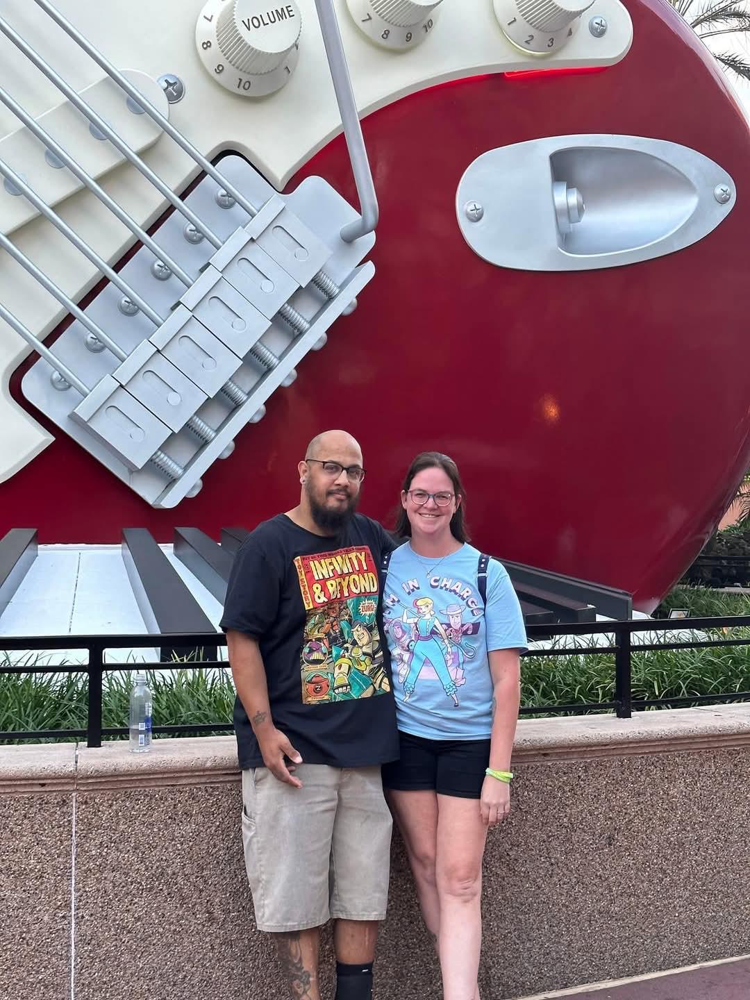 A couple smiles by a giant guitar statue at an outdoor entertainment venue.