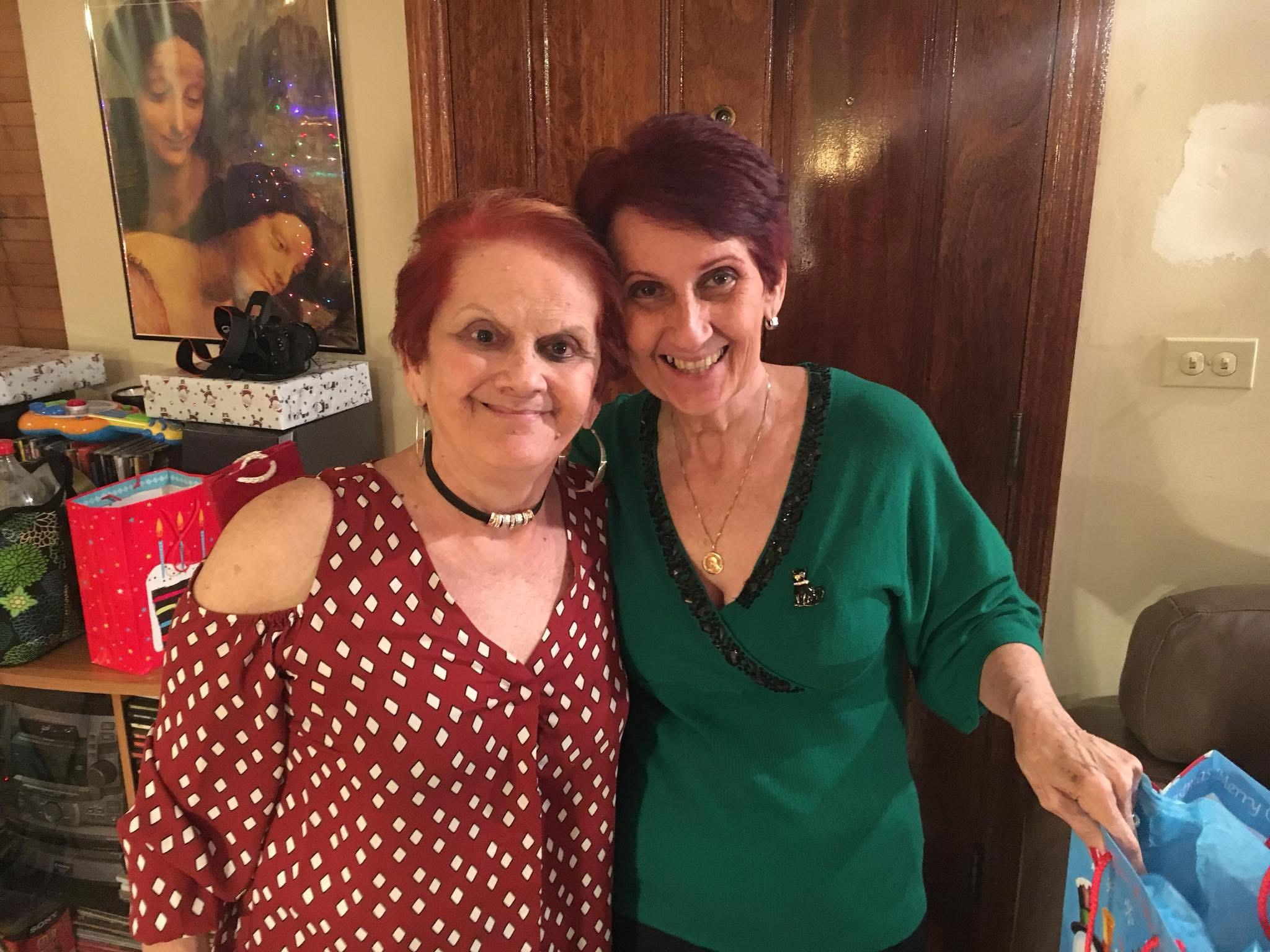 Two joyful women pose together with smiles in a festive living room filled with holiday decorations.