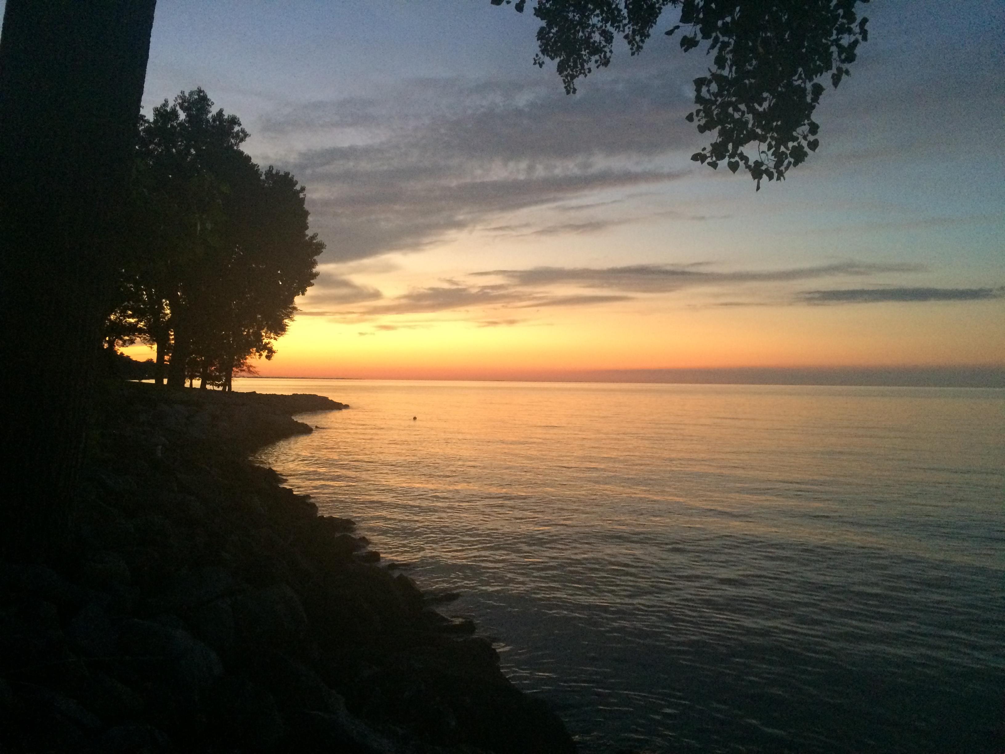 The sun sets over the lake, casting vibrant colors across the sky while trees stand silhouetted.