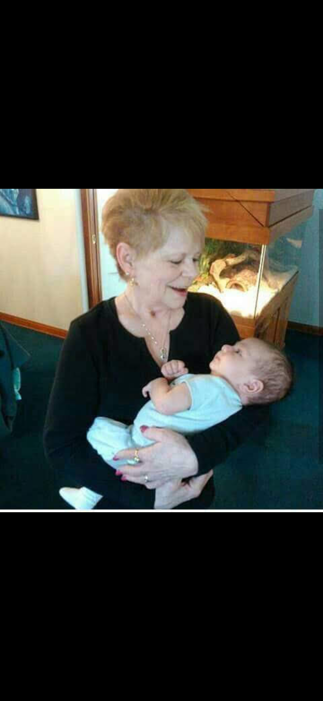 A grandmother smiles warmly at her newborn grandchild while cradling him gently in her arms.
