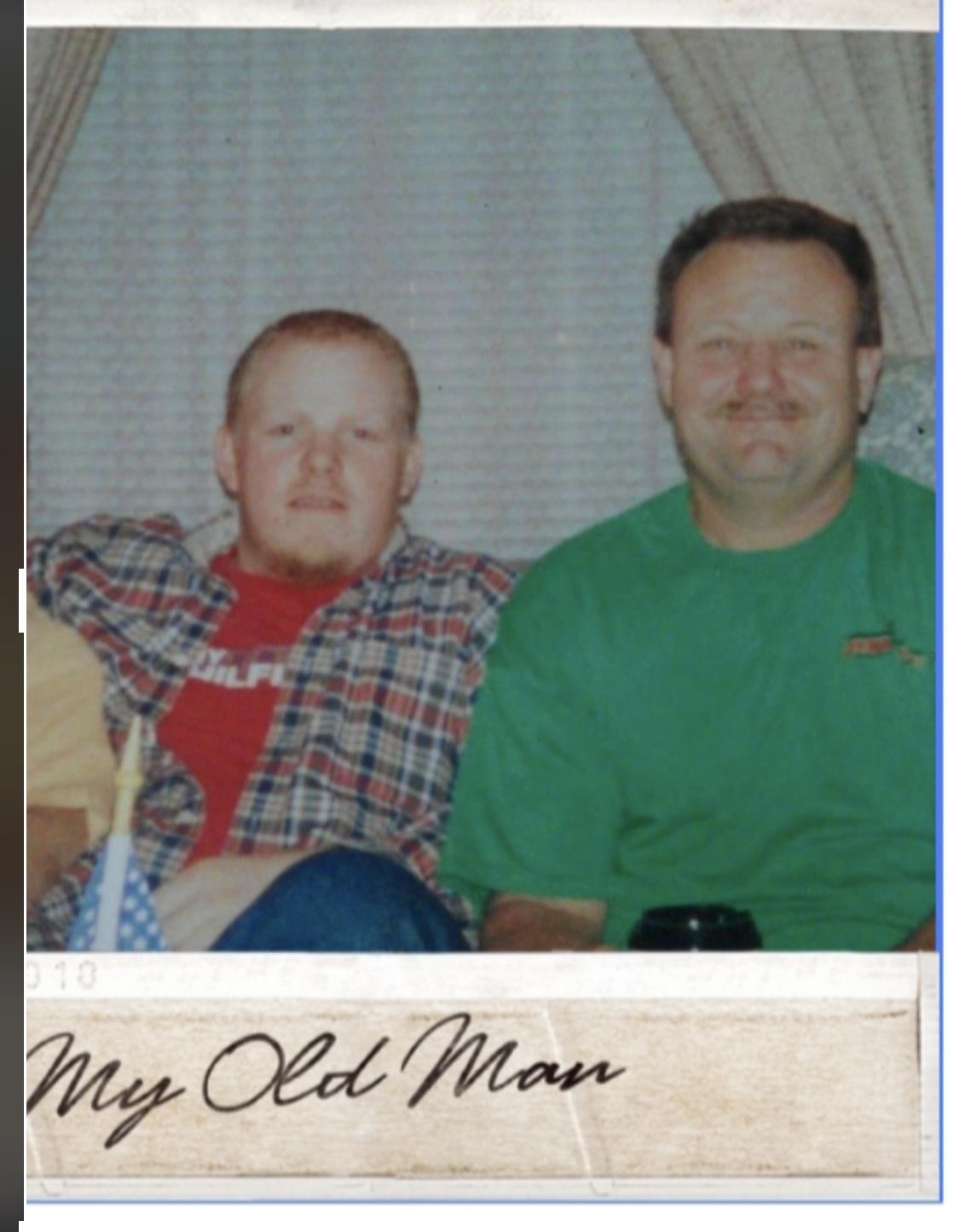 Two men are sitting on a couch, dressed casually and sharing a moment of camaraderie at home.