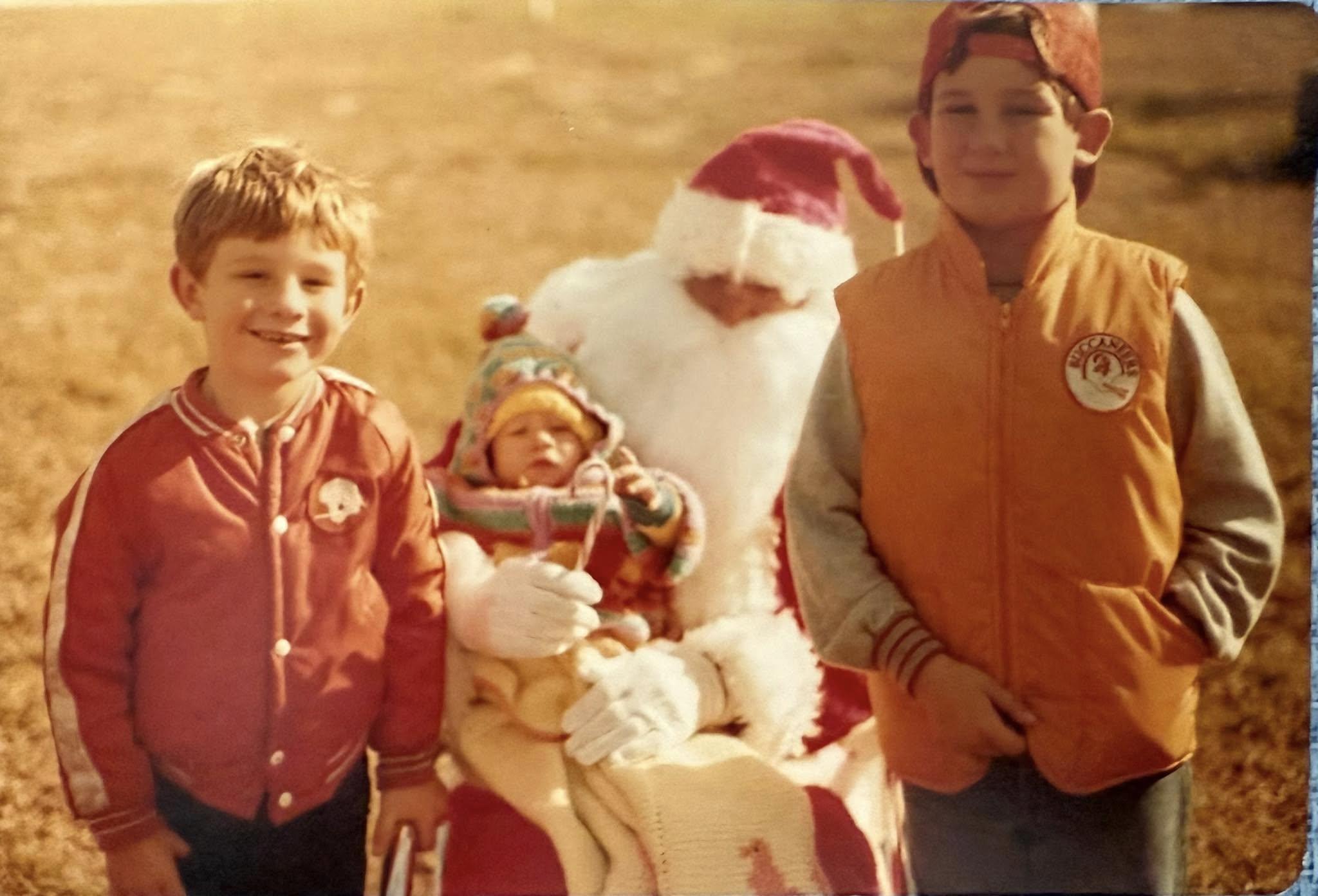 Two boys stand near Santa Claus, who holds a baby, all surrounded by grass on a sunny day.