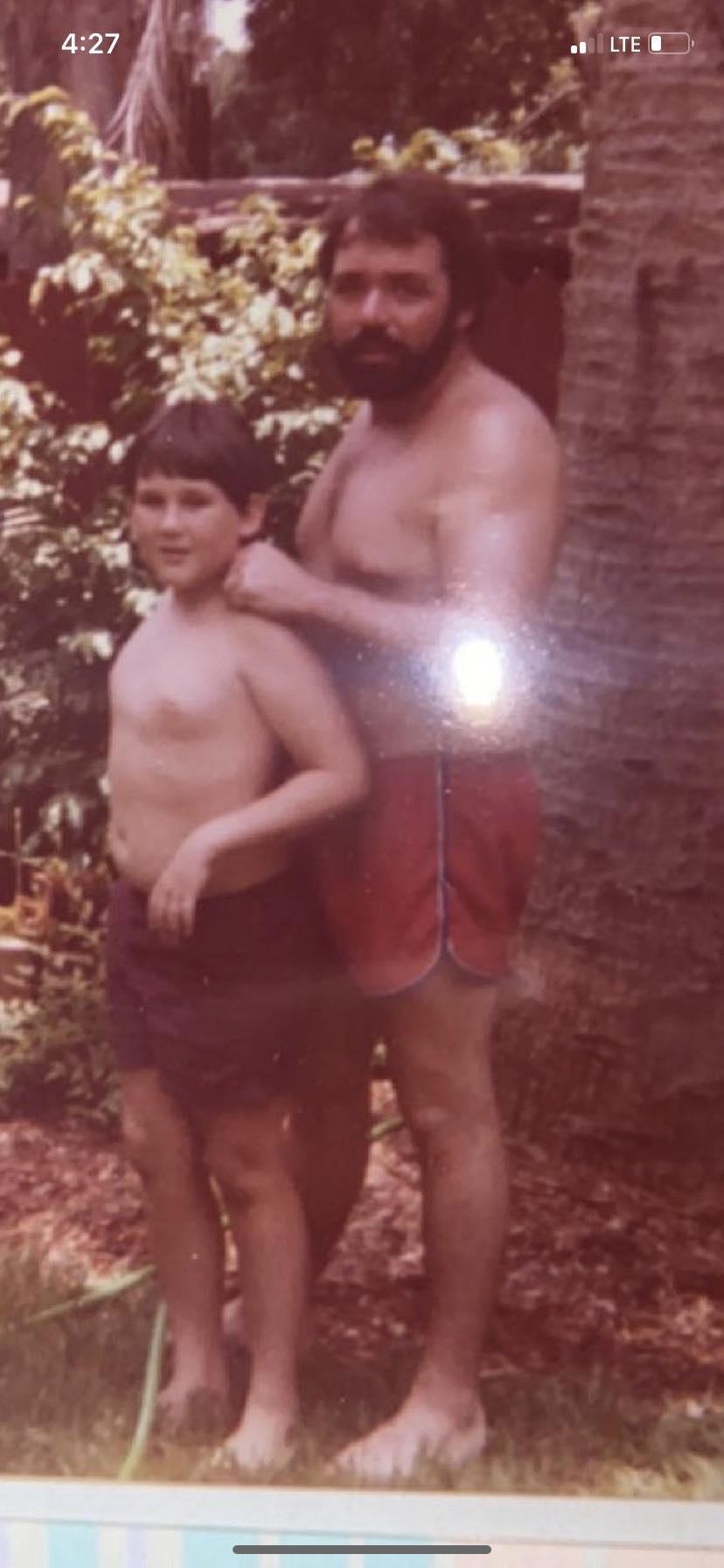 A shirtless father stands behind his son in a vibrant garden, both smiling and relaxed.