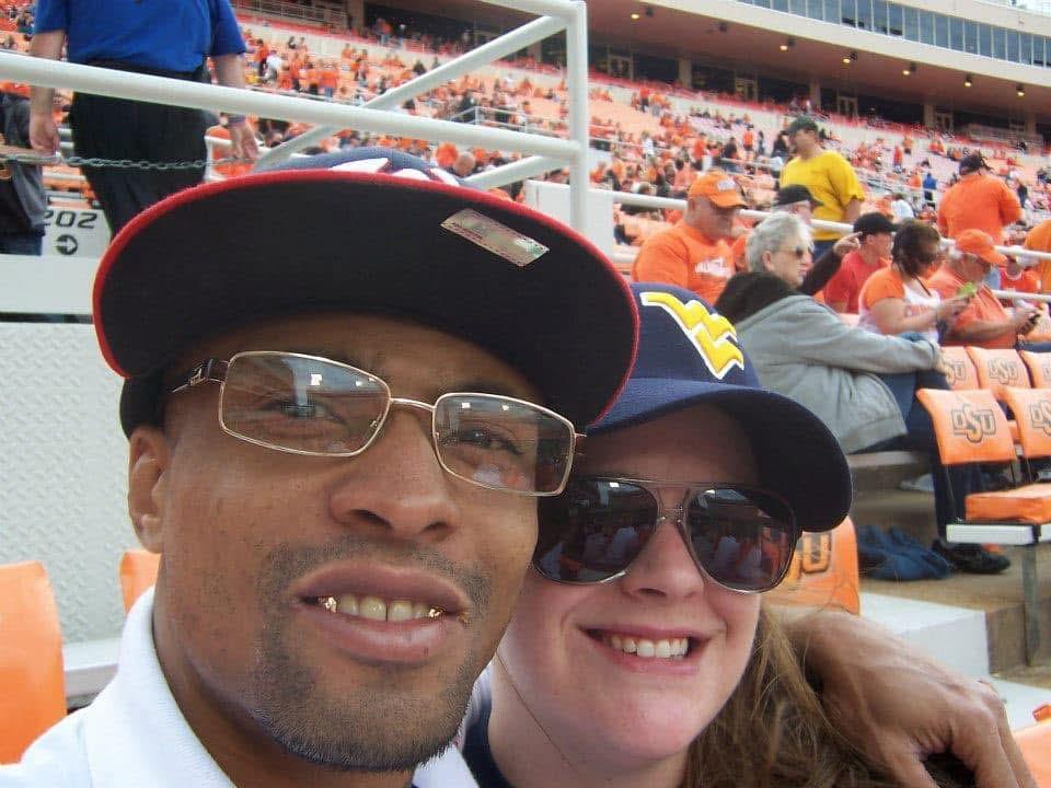 A couple smiles together at a college football game surrounded by cheering fans and excitement.