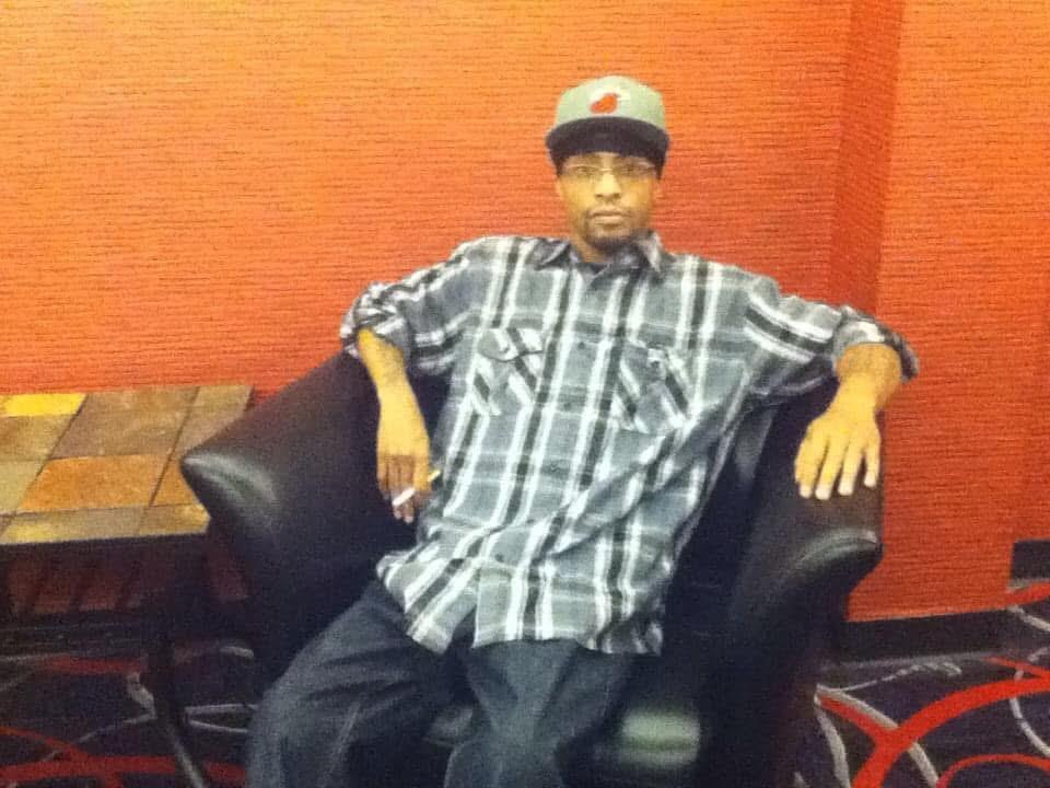 A man relaxes in a lounge chair wearing a plaid shirt and cap, showcasing a laid-back vibe.