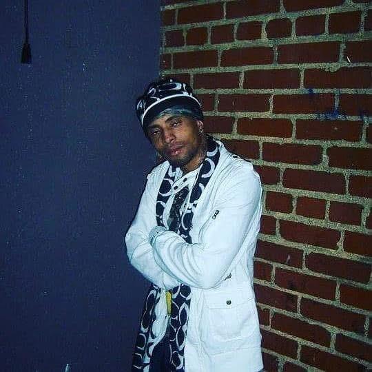 A man stands with crossed arms, showcasing an urban fashion look against a brick wall backdrop.