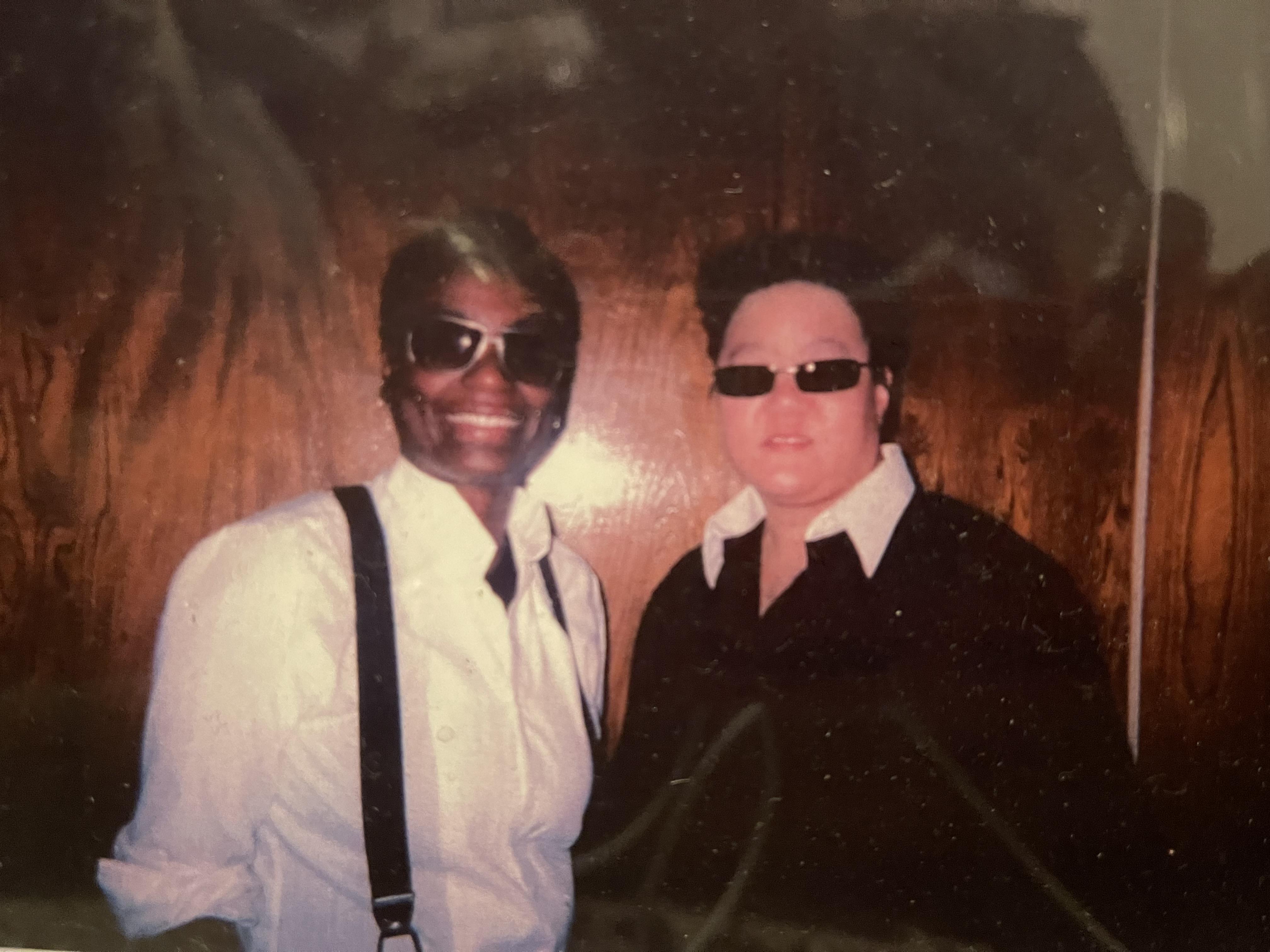 Two people with sunglasses stand close together, smiling at a social gathering indoors.