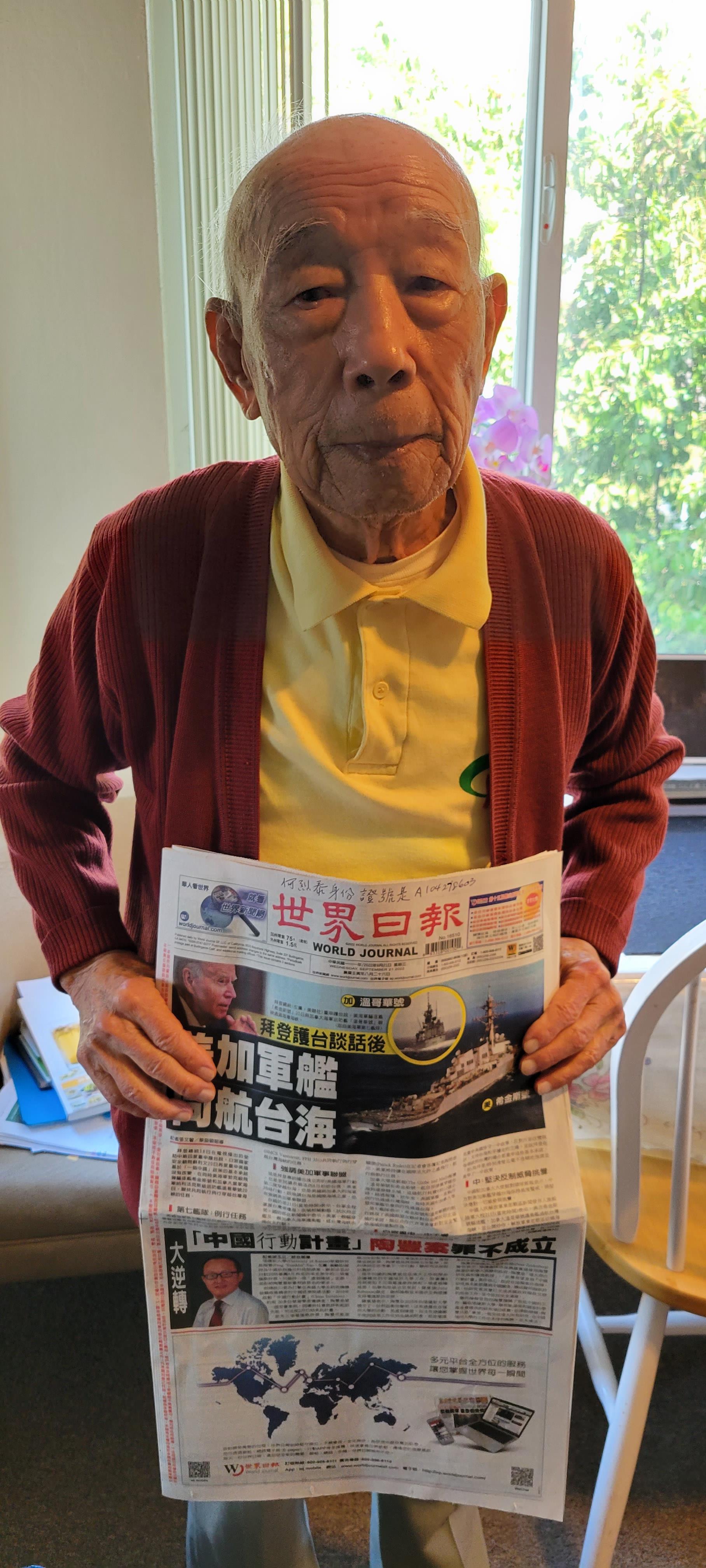 An elderly man with gray hair stands in a well-lit room holding a newspaper, sharing news.