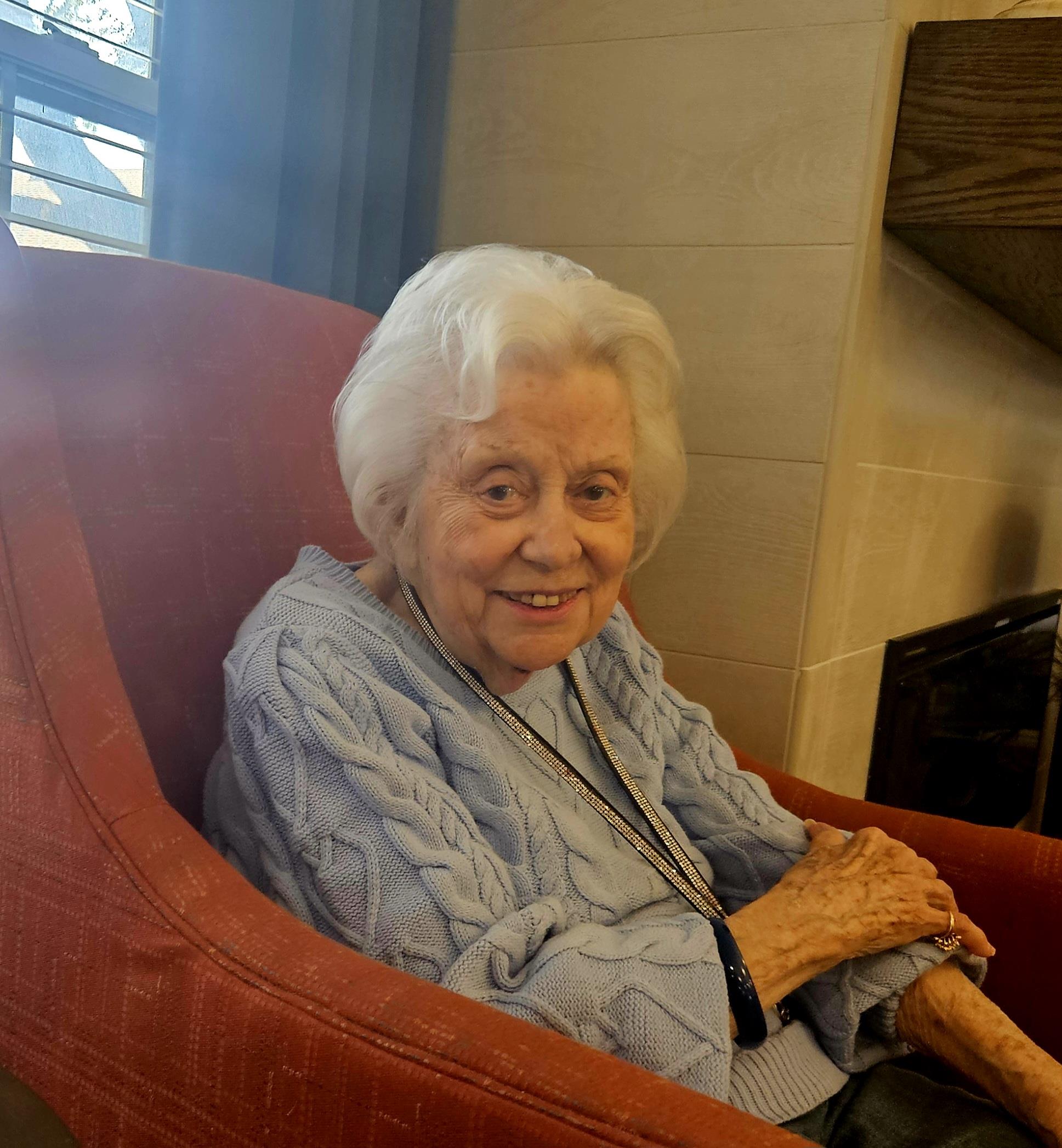 An elderly woman with white hair sits happily in a cozy chair, enjoying her surroundings.