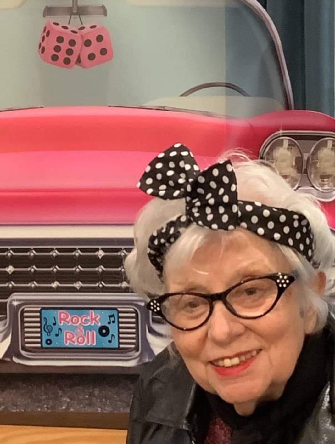 Elderly woman with stylish bow enjoying a retro-themed event near a pink vintage car.