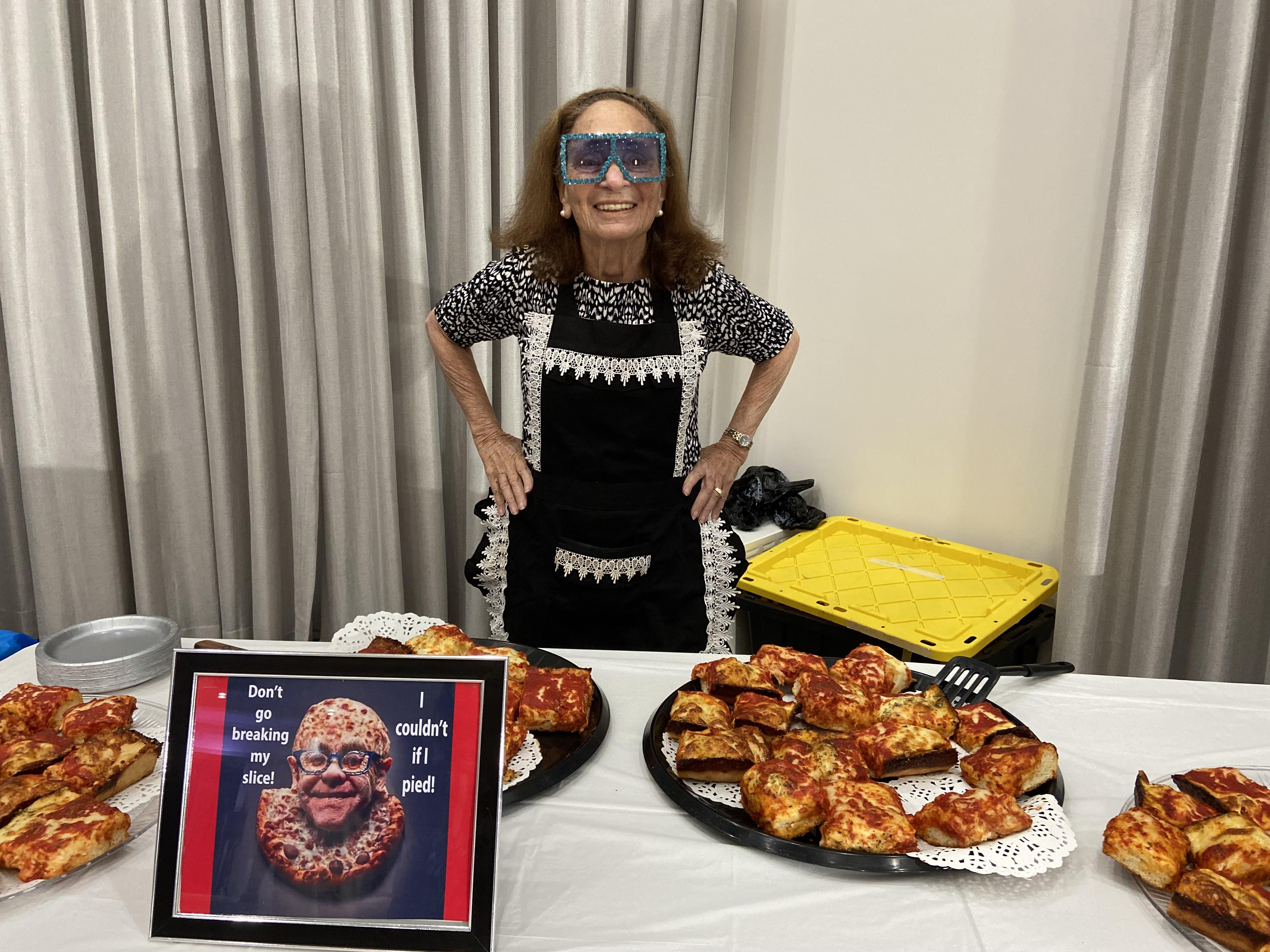 An elderly woman with playful glasses showcases a variety of pizzas at a local gathering.