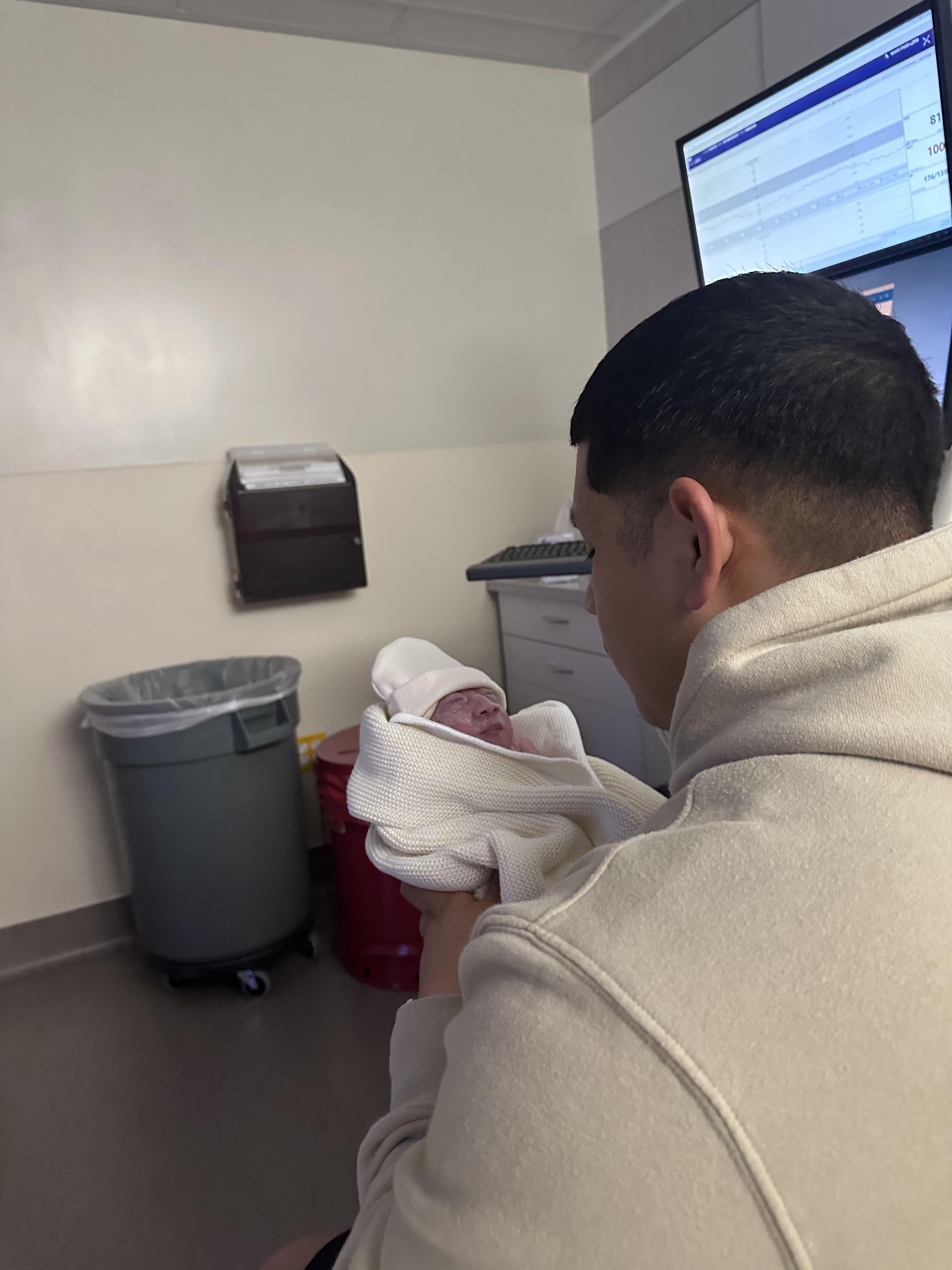 A father cradles his newborn baby wrapped in a blanket in a hospital setting.